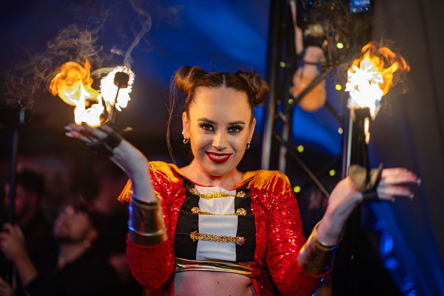 A performer in a red sequined costume holds flaming torches during a nighttime show.