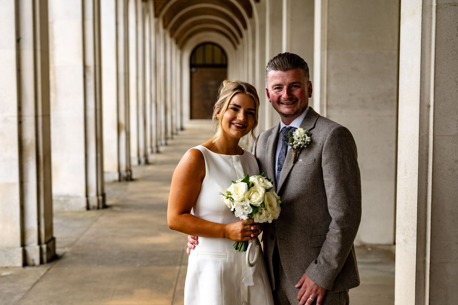 A couple shares a romantic moment in a colonnade walkway with white stone arches and natural lighting.