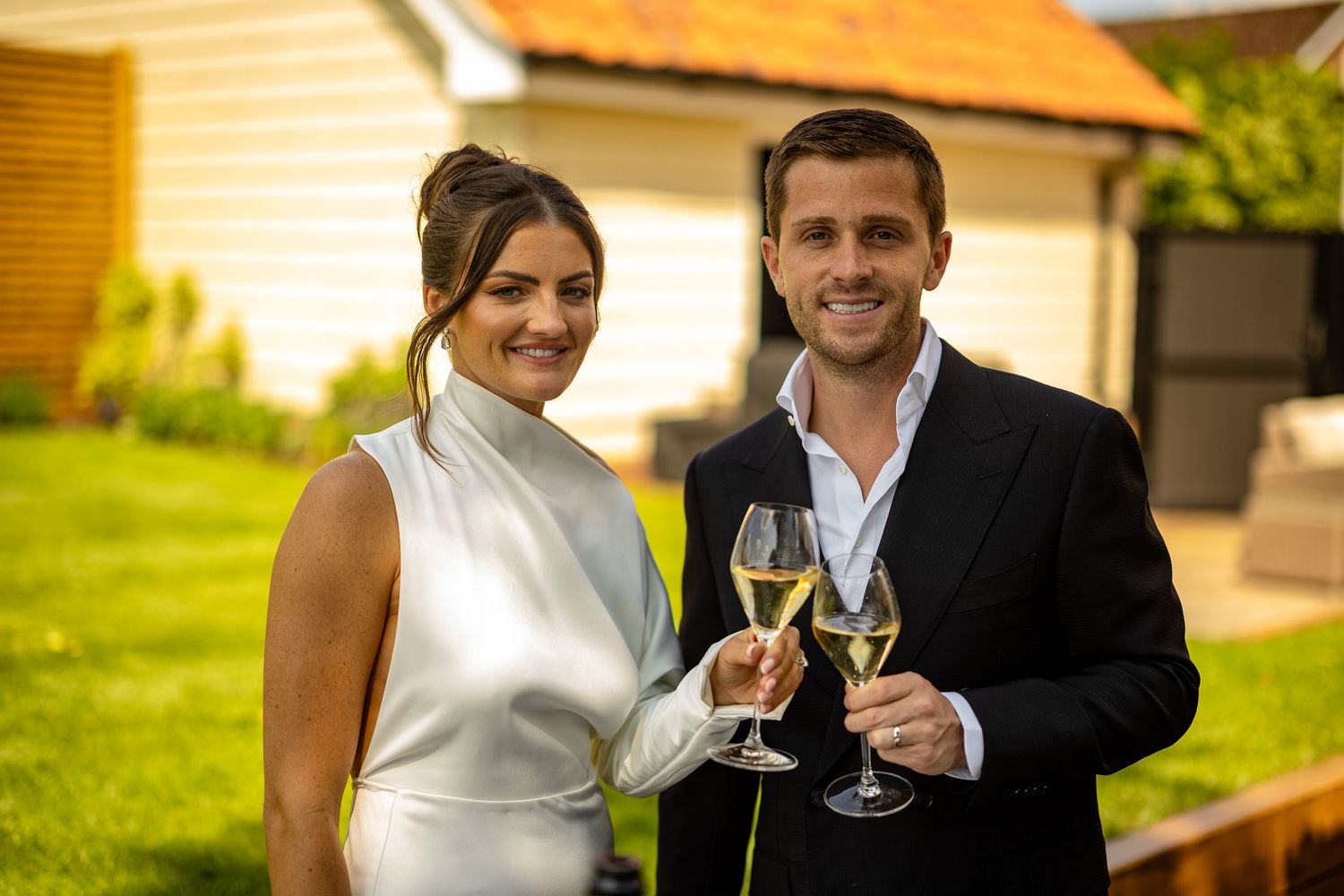 A couple in formal attire toasts with wine glasses while enjoying an outdoor celebration.