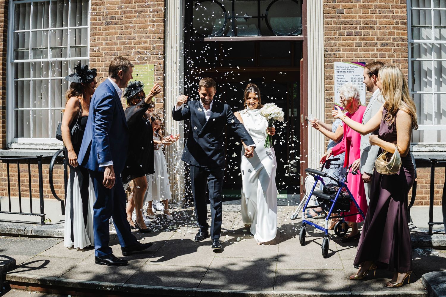 Wedding celebration outside a brick building with guests throwing confetti.
