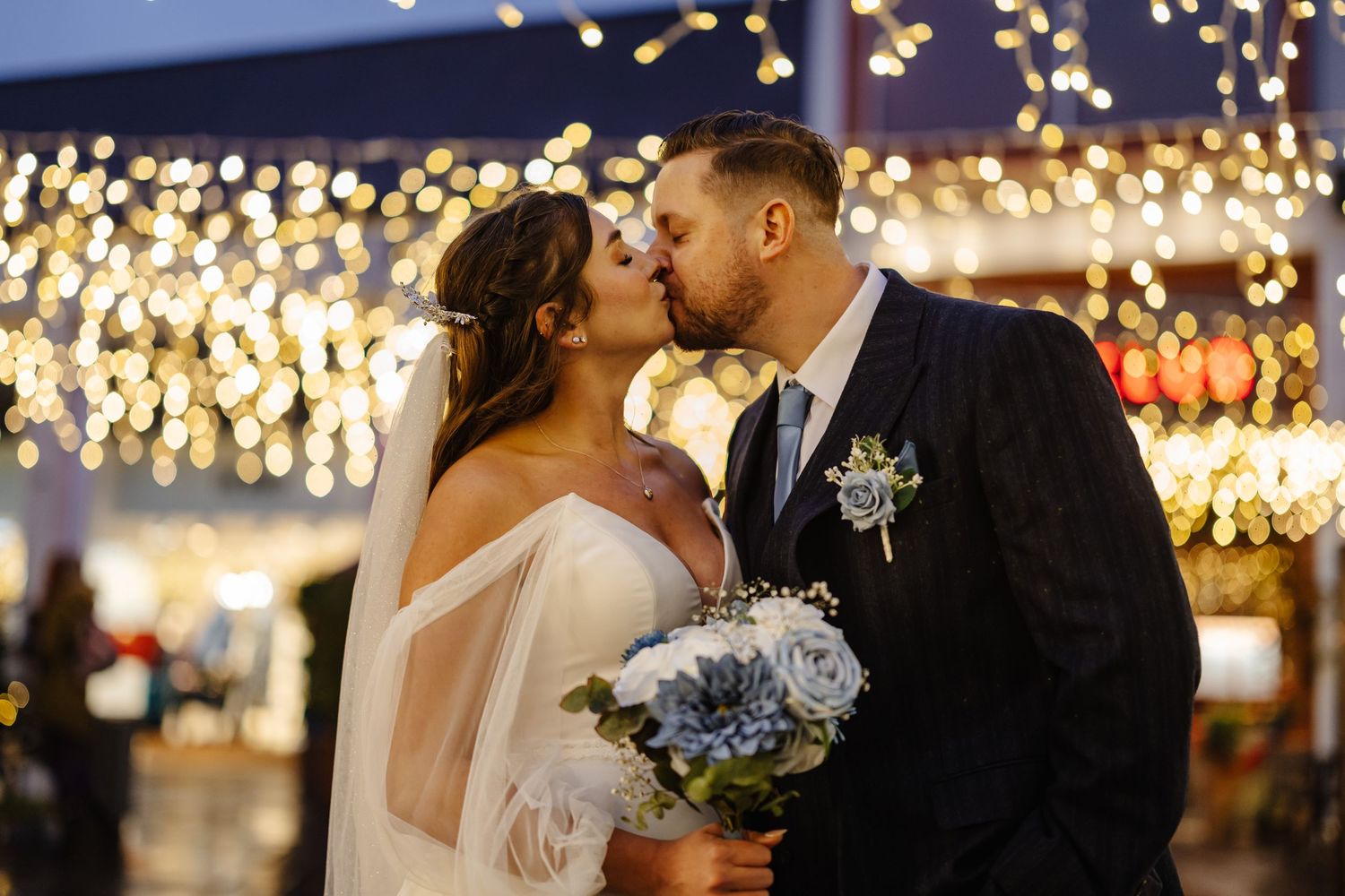 A romantic wedding couple shares a kiss under twinkling string lights while the bride holds a blue and white bouquet.