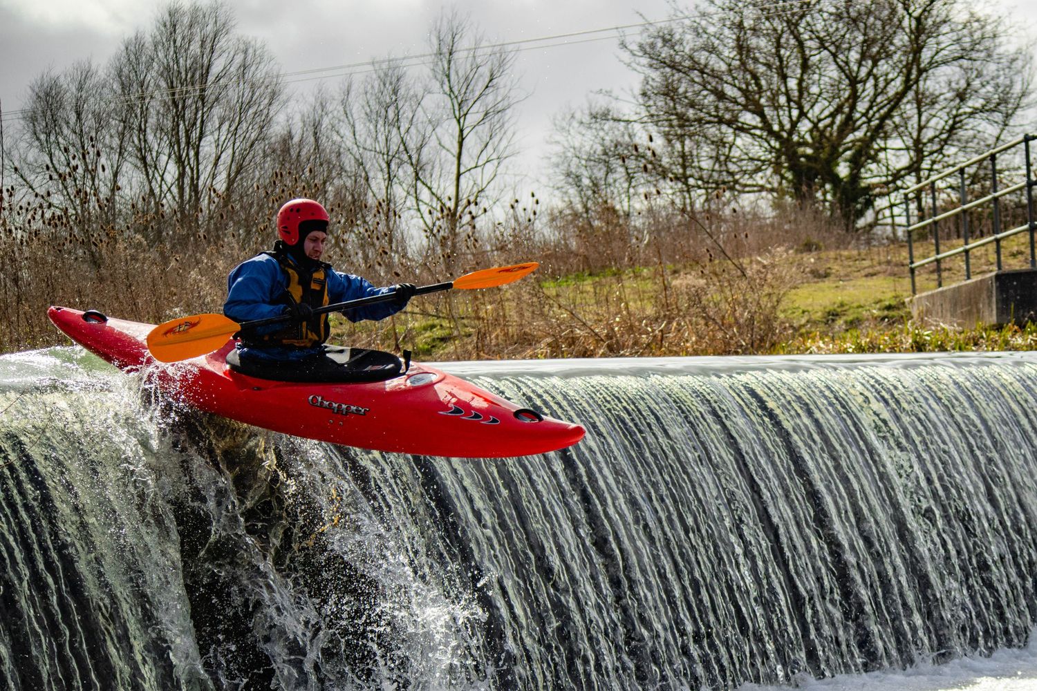A kayaker navigates over a small waterfall dam in a river during autumn season.