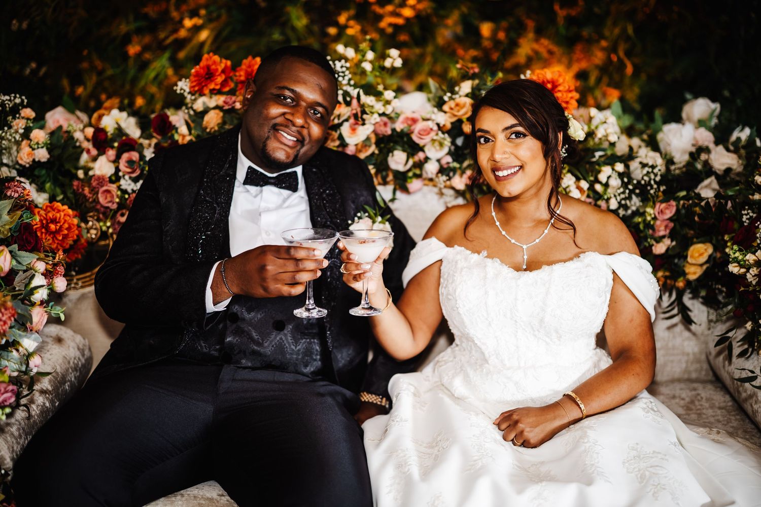 A newlywed couple shares a toast while sitting together surrounded by colorful floral arrangements at their wedding.