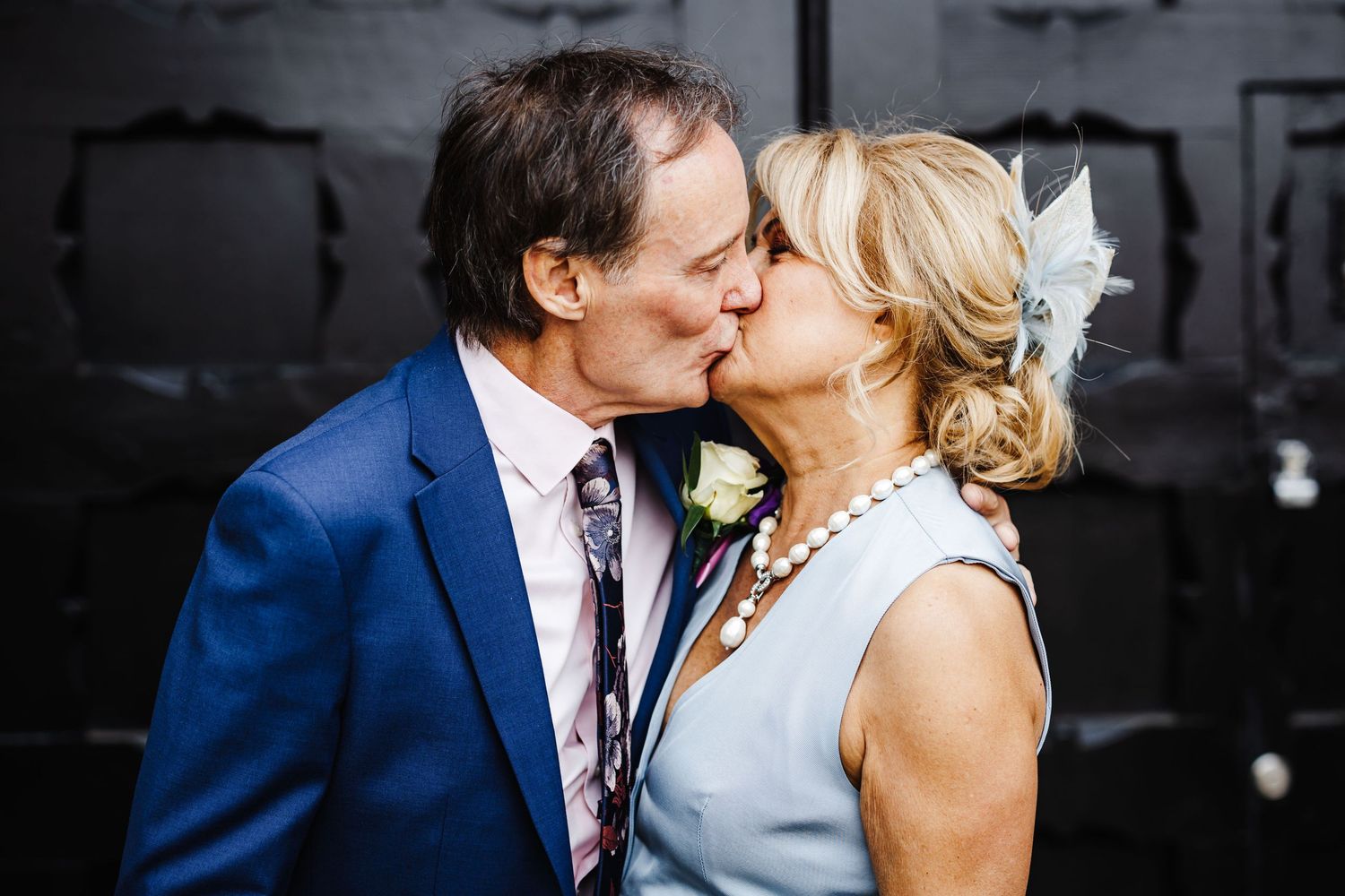 A couple shares a romantic kiss while dressed in formal attire, with the person on the left wearing a blue suit.