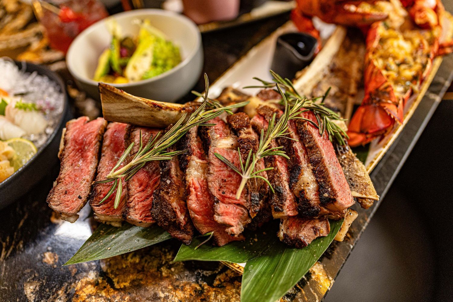 Sliced medium-rare ribeye steak garnished with fresh rosemary sprigs and served on a dark platter with grilled lobster.