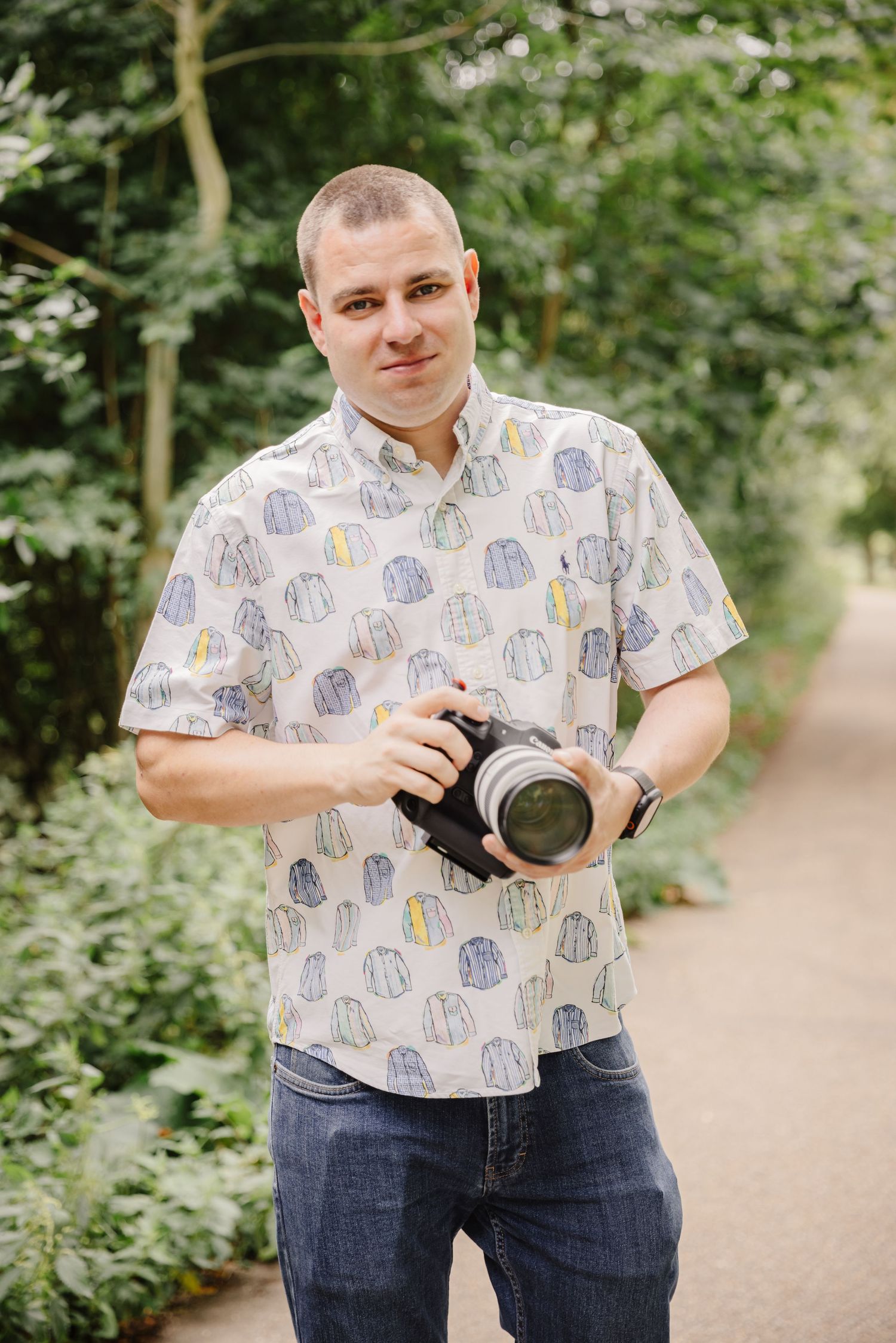 A photographer in a patterned shirt holding a camera on a wooded path.
