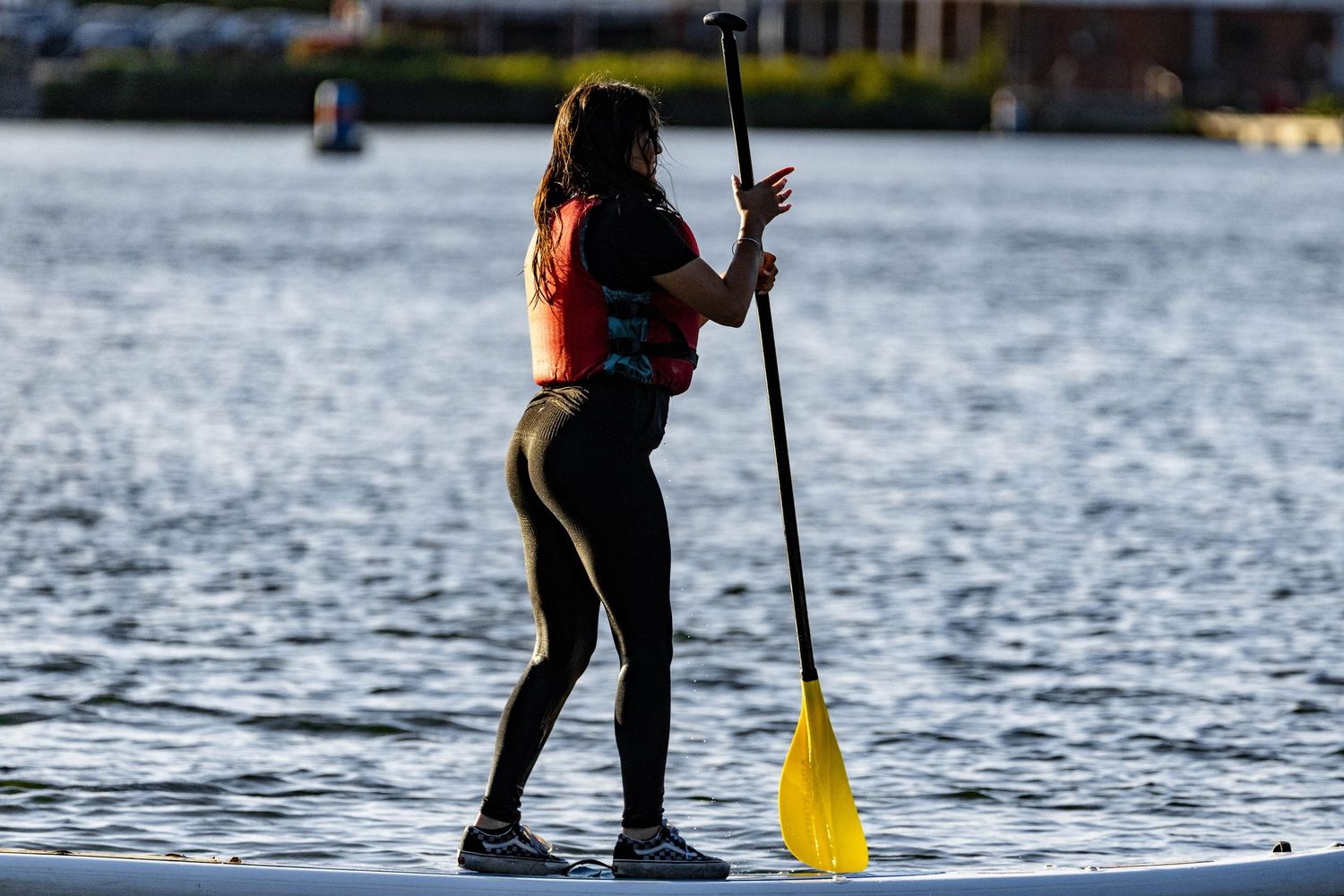 A paddleboarder in an orange life vest stands on calm waters holding a yellow paddle at sunset.