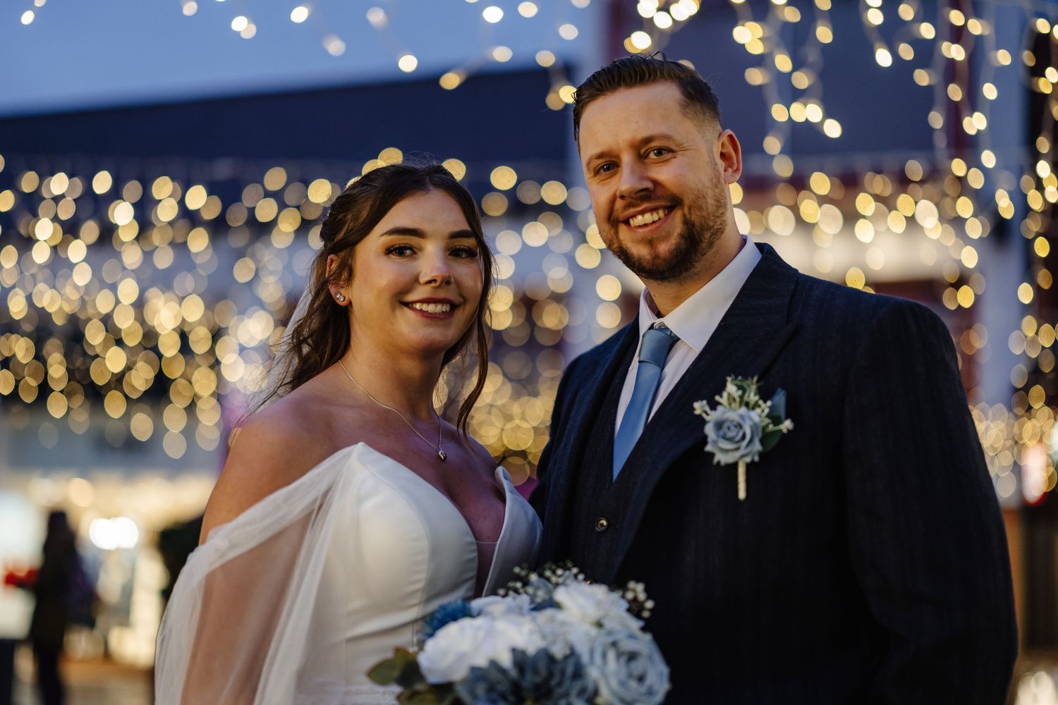 A couple shares a joyful moment under twinkling string lights at an evening wedding celebration.