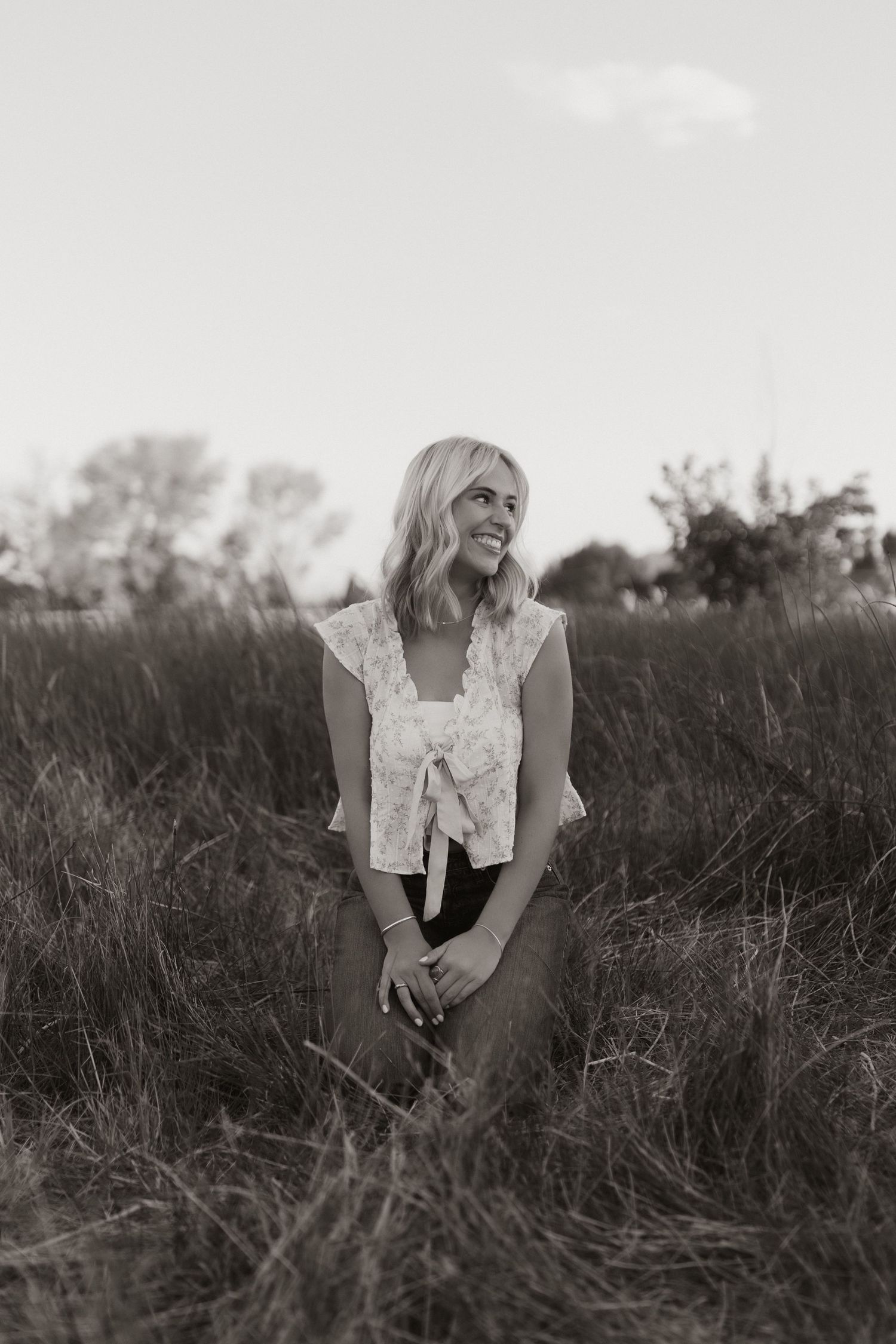 A person in a white ruffled top sits among tall grass in a dreamy black and white portrait.