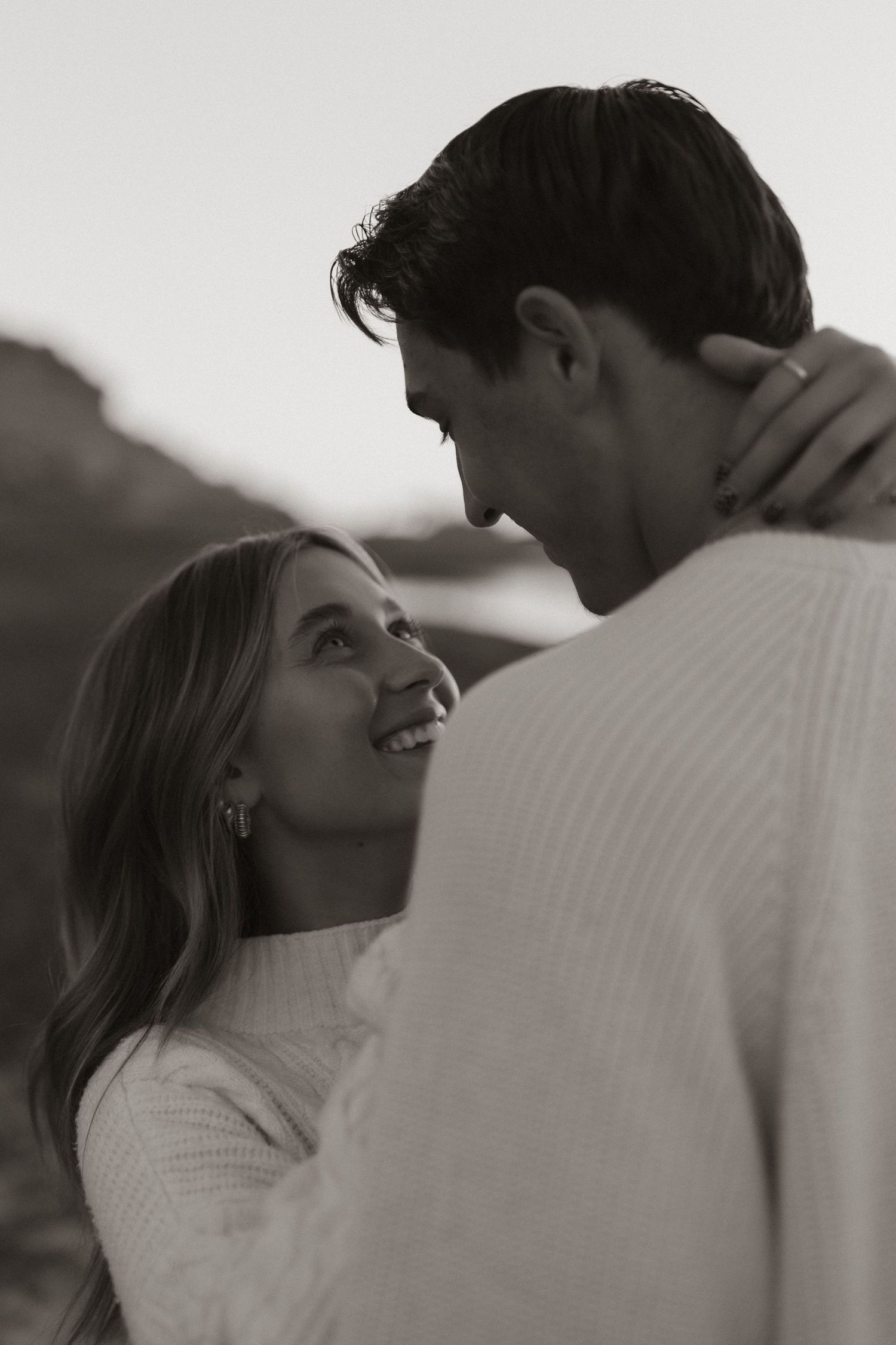 A romantic black and white portrait of a couple in white sweaters gazing at each other with genuine smiles.