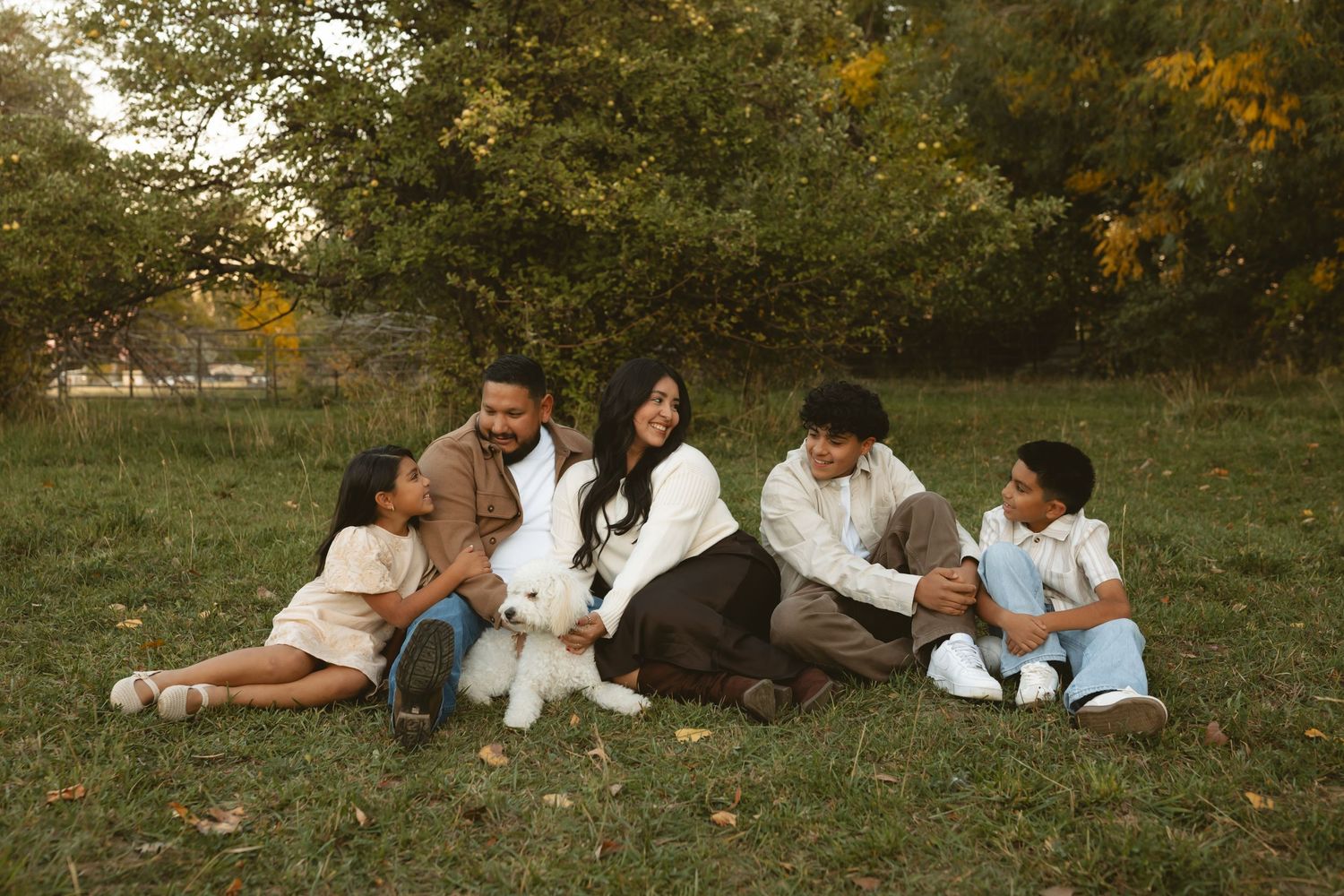 A family sits together on grass during an autumn day, wearing coordinated casual outfits in neutral tones.