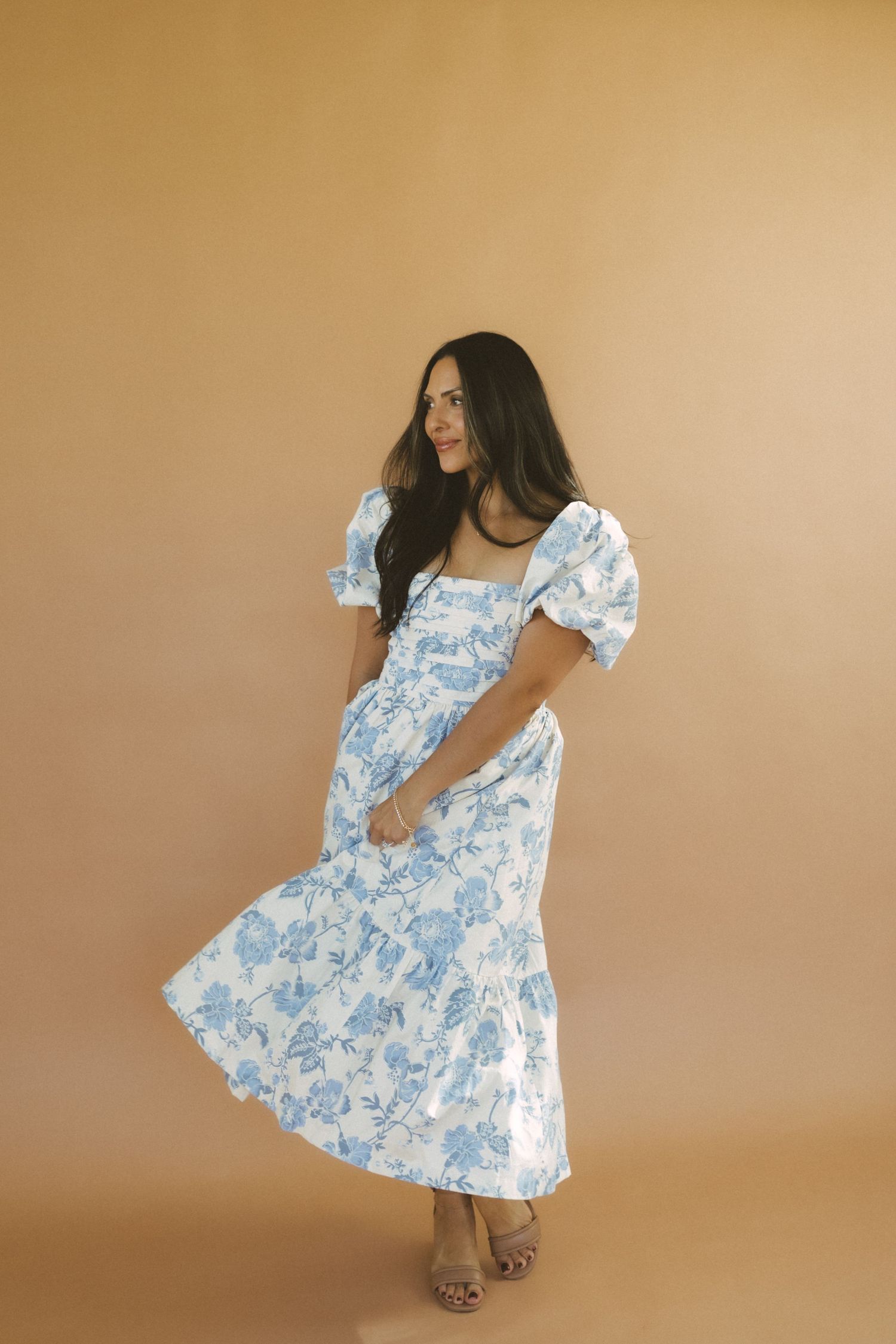 A person poses in a flowing white and blue floral print dress against a neutral beige backdrop.