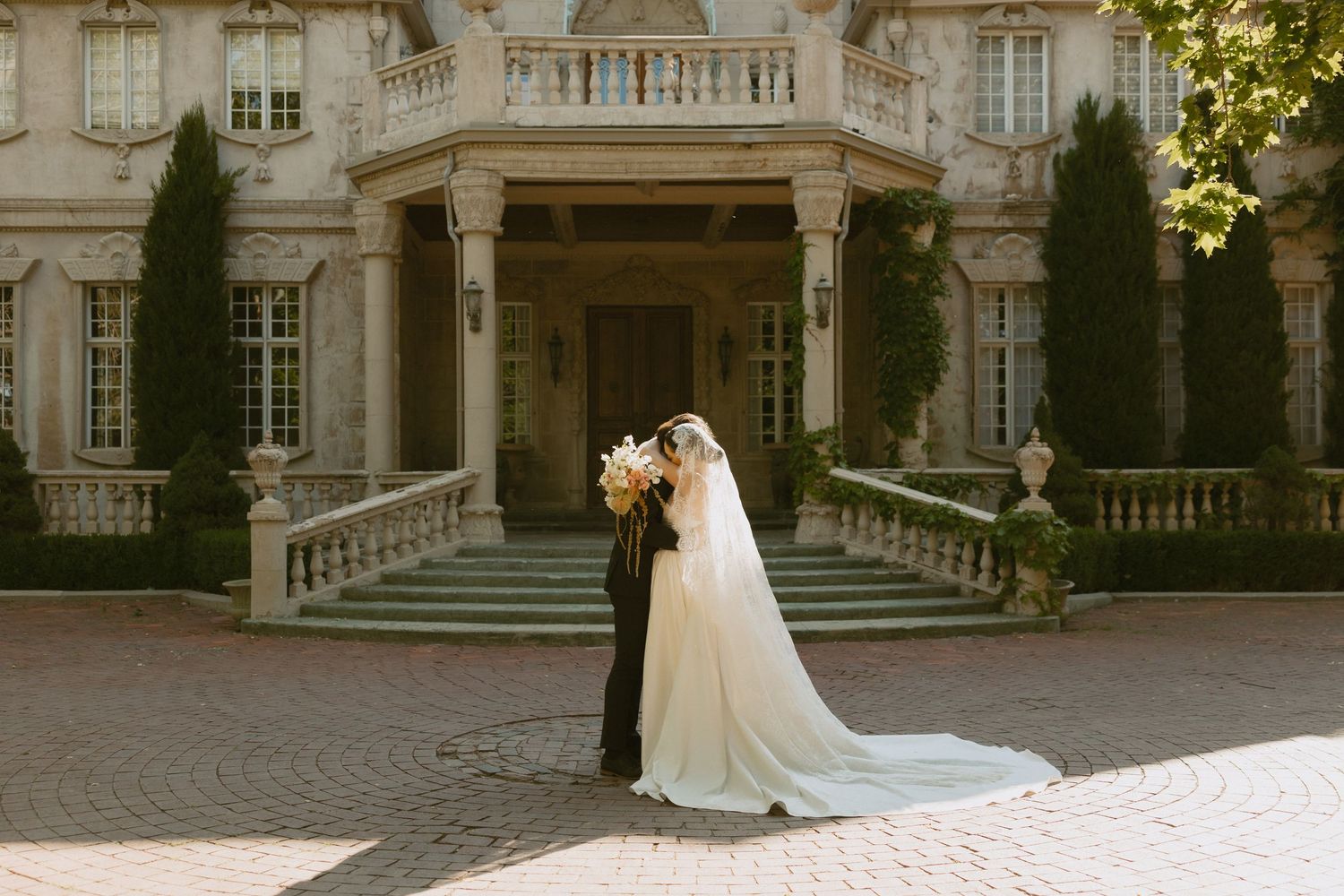 A romantic wedding scene at a grand mansion entrance with stone steps and elegant architecture in warm sunlight.
