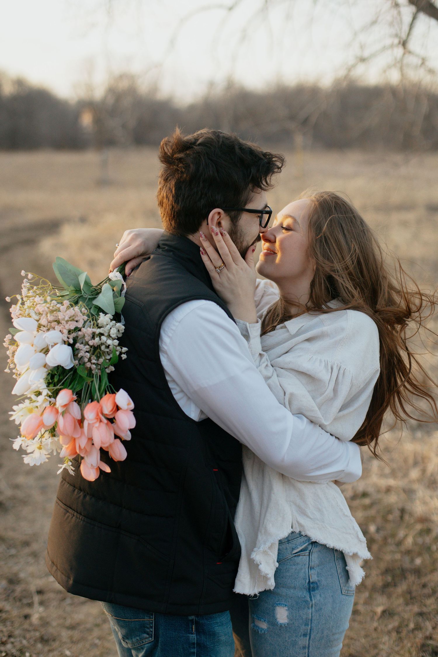 Romantic couple kissing outdoors holding pink tulip bouquet in a field during golden hour sunset.