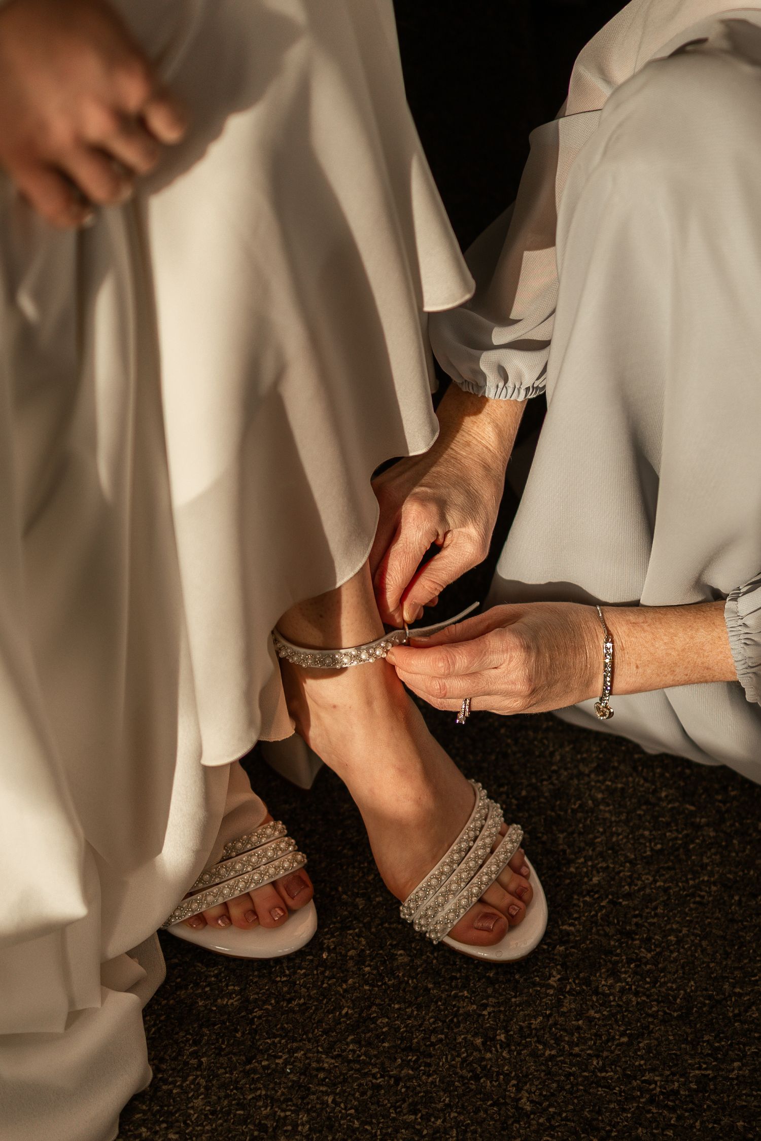 Hands fastening sparkly sandal straps on a carpeted floor with white fabric flowing in the background.