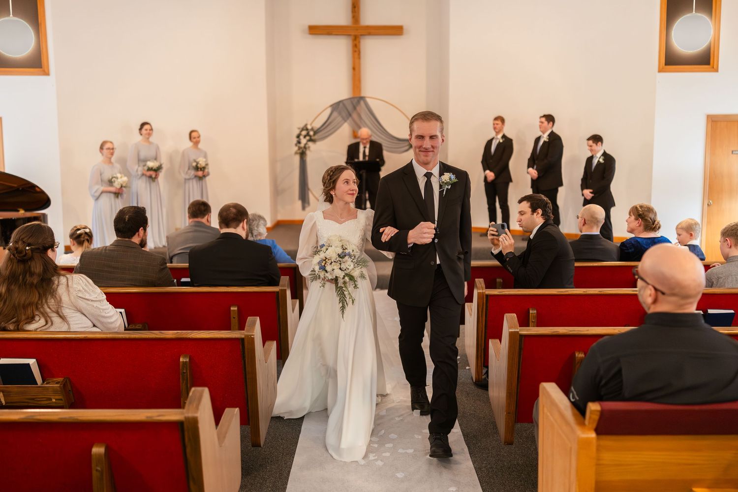 A couple walks down the aisle of a small chapel with red pews and a wooden cross at the altar during a wedding ceremony.