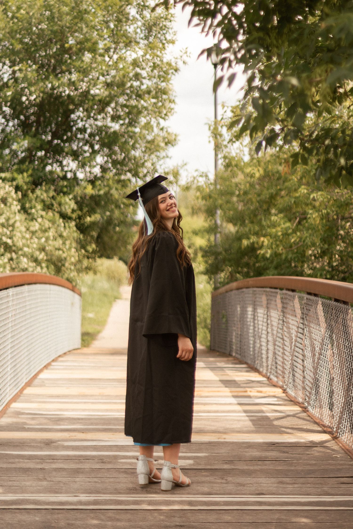 A person in graduation attire walking across a wooden bridge surrounded by trees.