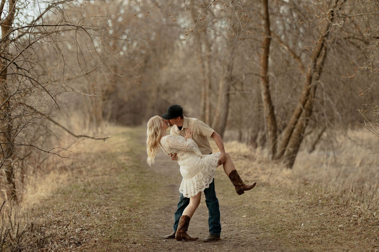 Couple sharing a romantic dip kiss on a wooded path, wearing cowboy boots and a white lace dress in autumn.