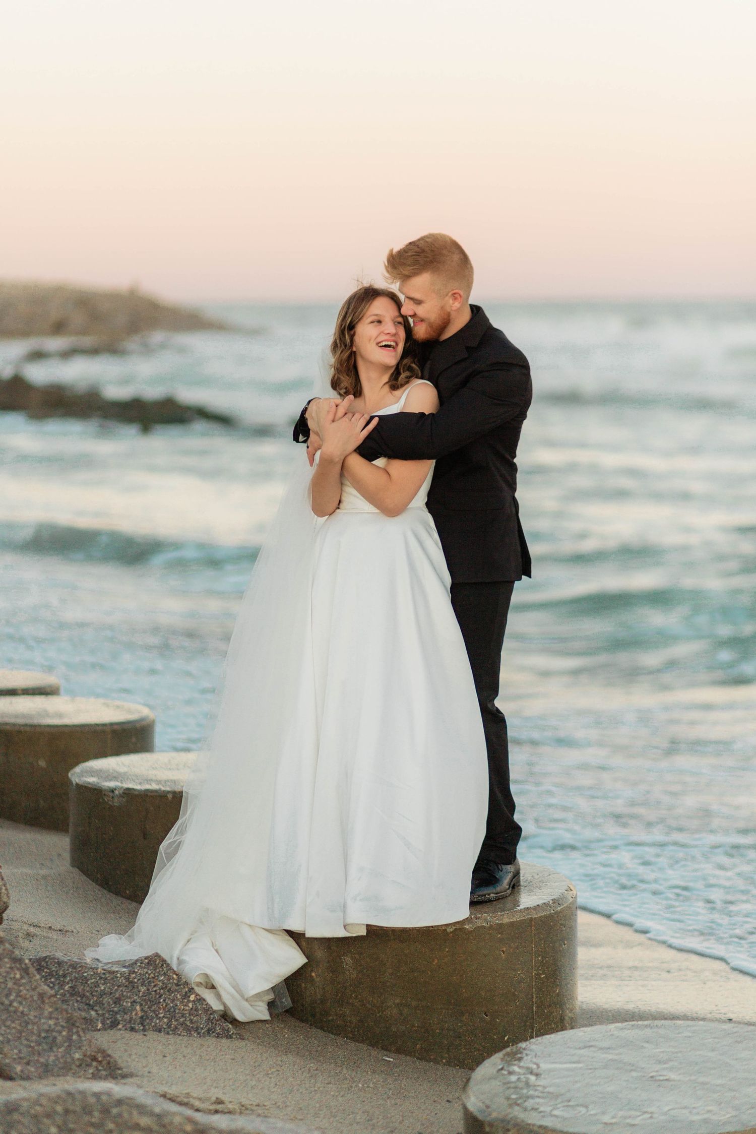 A romantic sunset beach wedding photo with a couple embracing on concrete steps by the ocean waves.