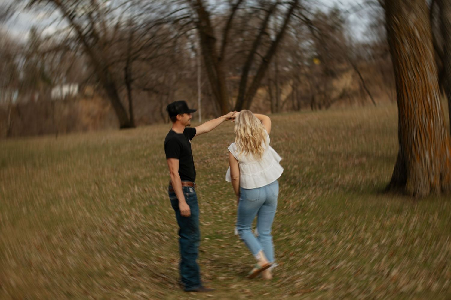 Couple dancing in a field with bare trees, woman in white top and jeans, man in black shirt and cowboy hat.