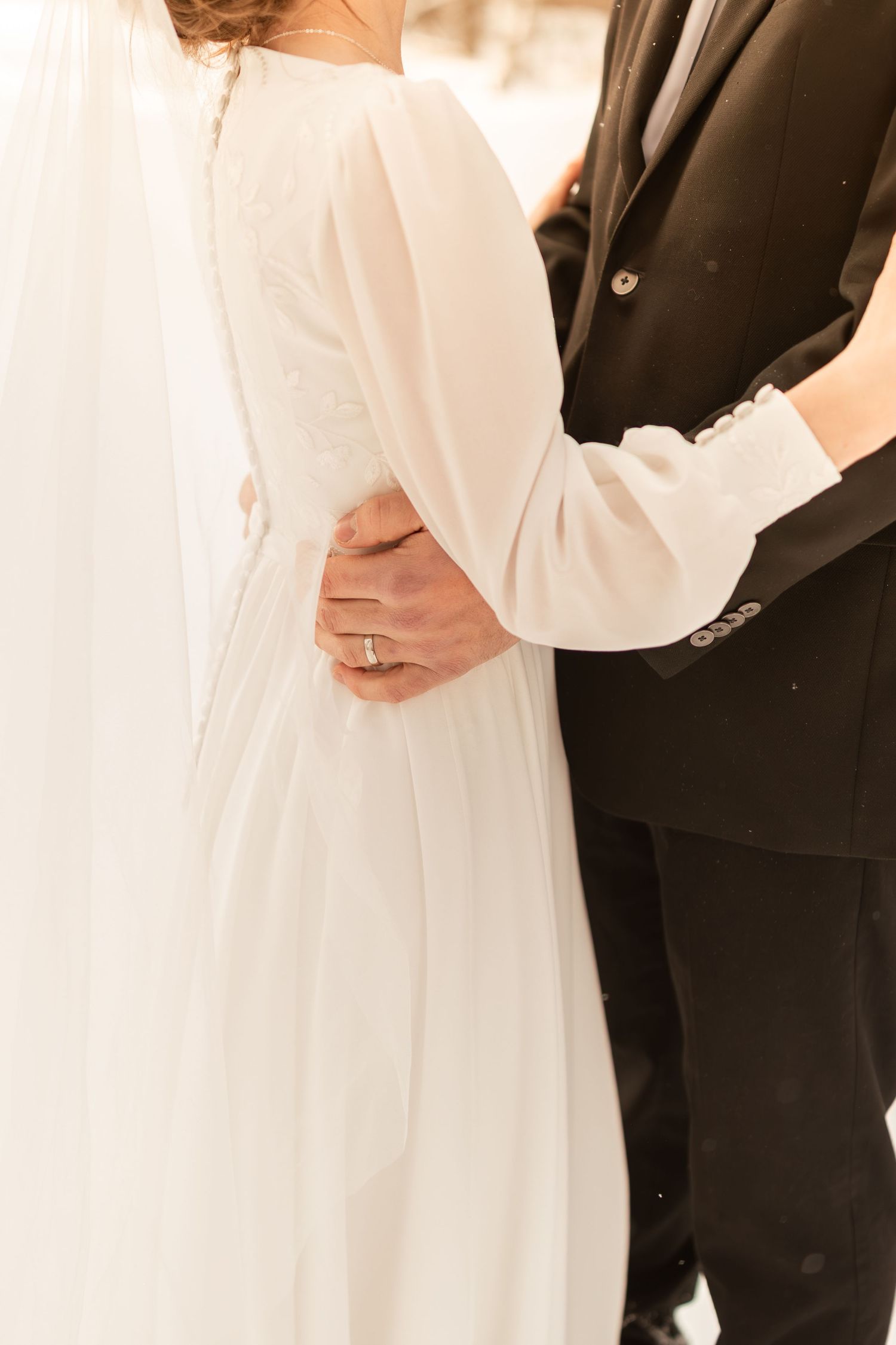 A couple in formal wedding attire embrace, with the bride wearing a flowing white dress and groom in classic black suit.