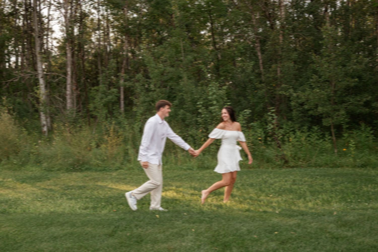 A couple in white outfits dances playfully in a lush green outdoor setting.