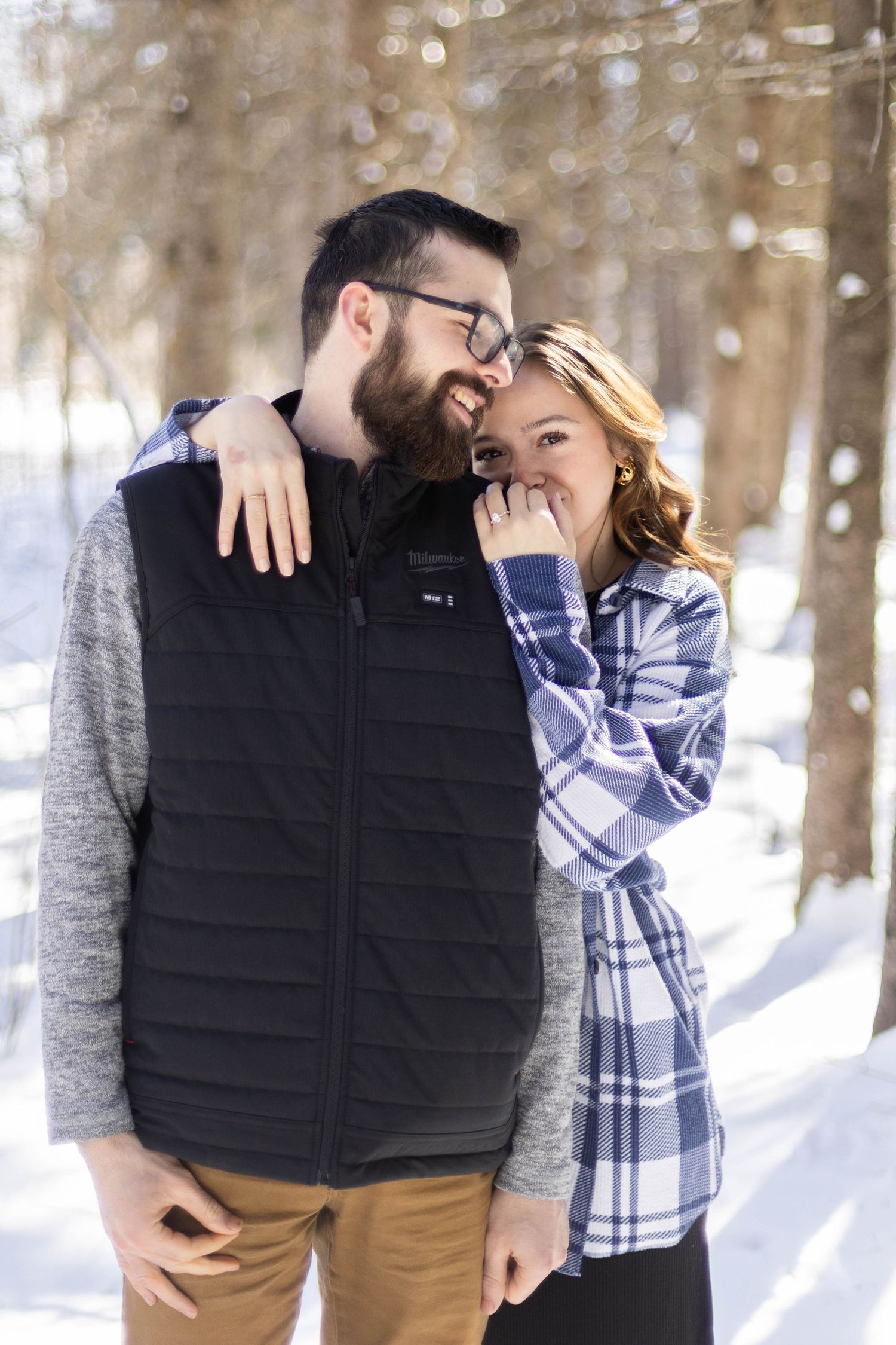 A couple embraces in a snowy winter forest setting wearing casual clothing.