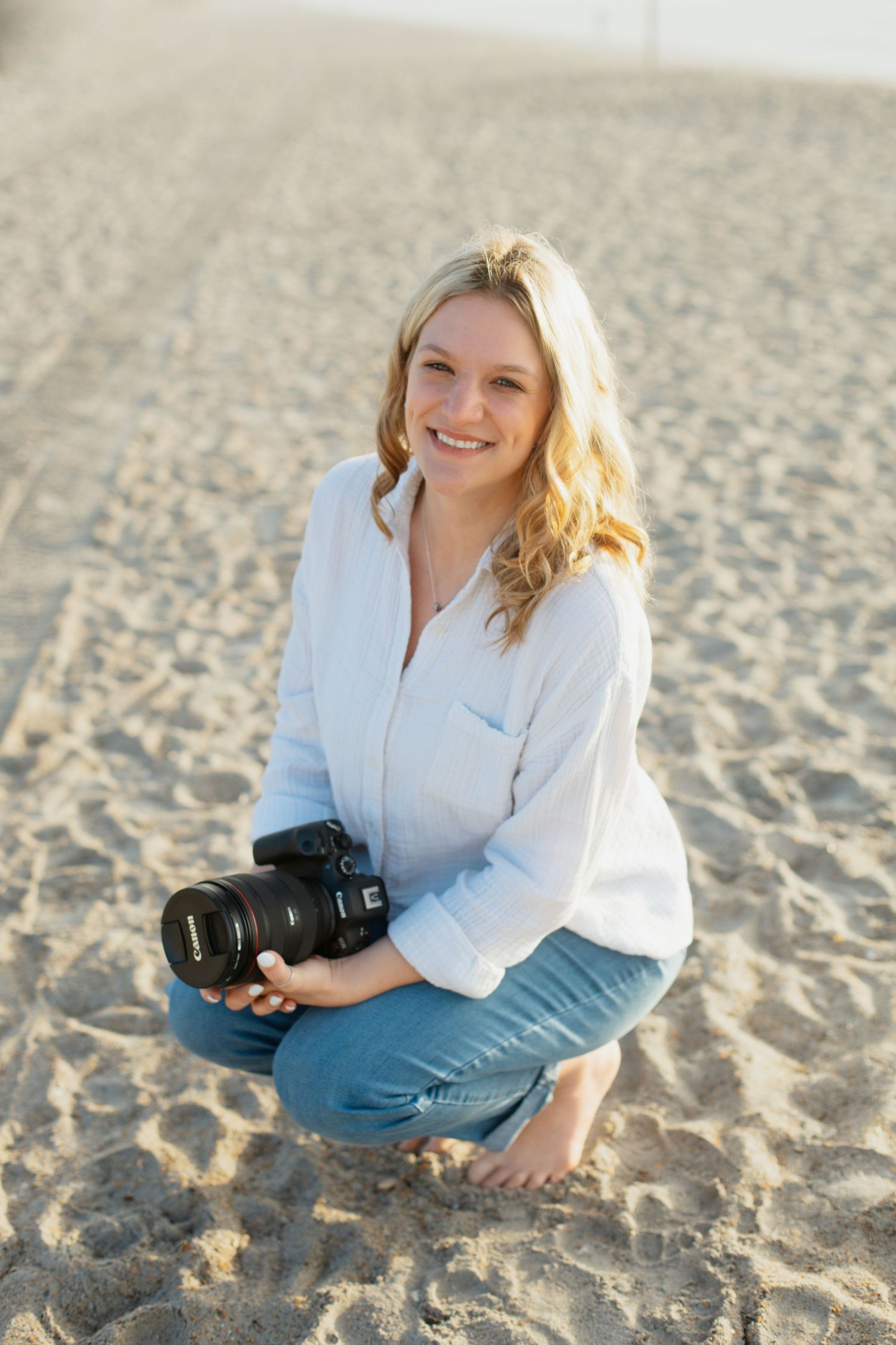 Smiling blonde woman crouching on sandy beach holding a Canon DSLR camera, wearing white shirt and jeans.