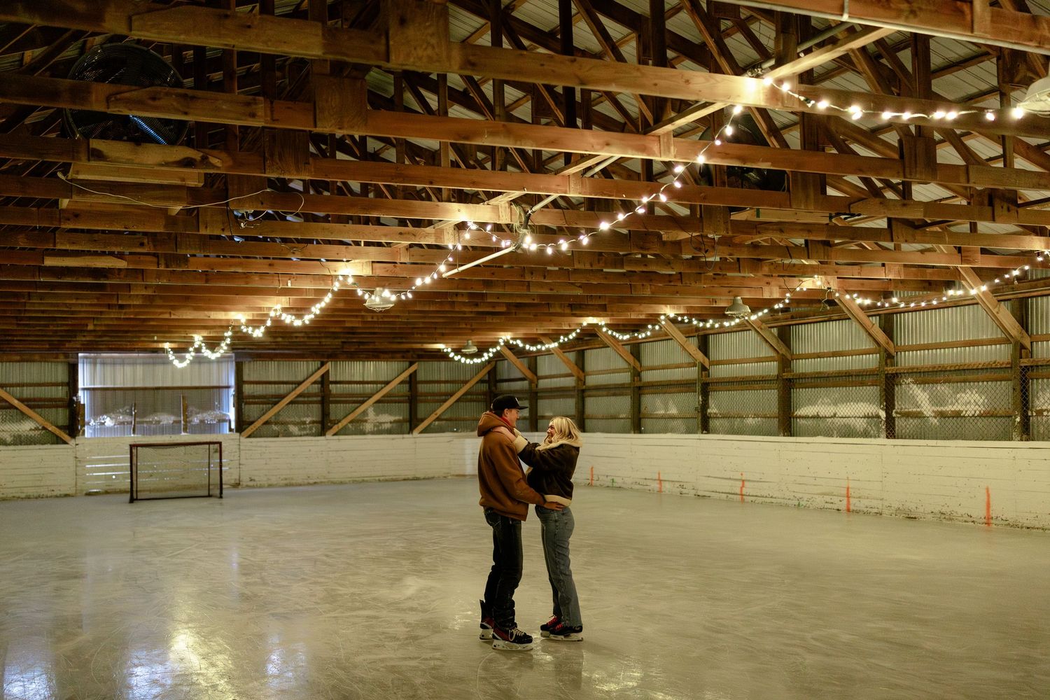 People skating on an indoor ice rink illuminated by string lights hanging from wooden rafters above.
