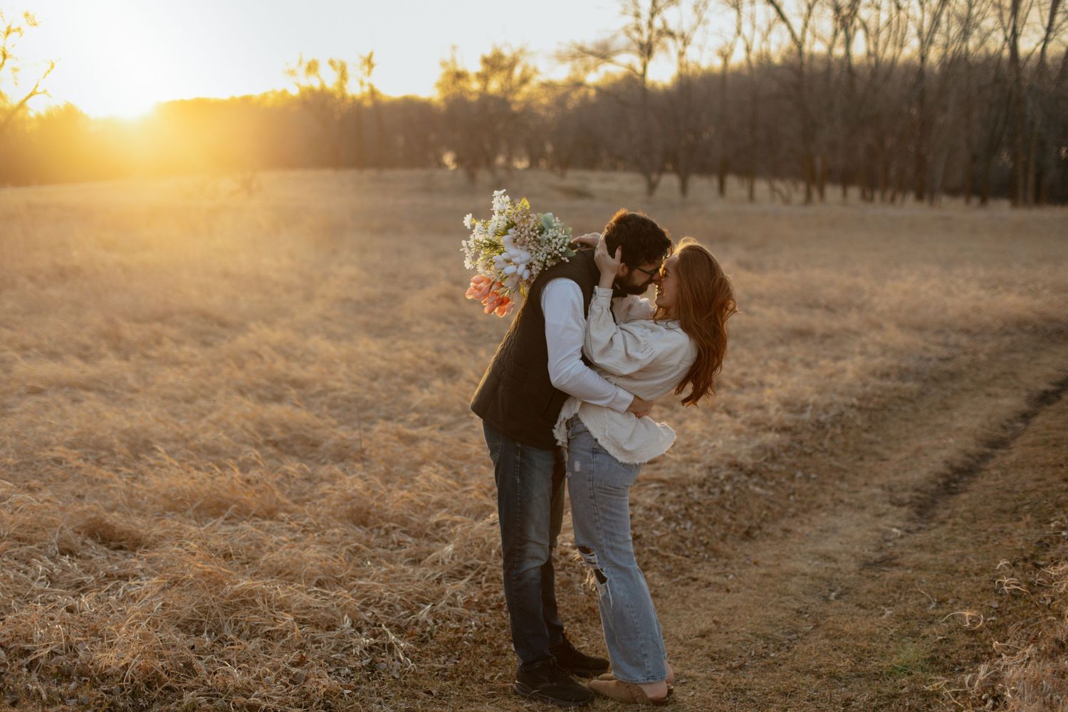 Couple sharing a romantic kiss at golden hour in a field, man holding a wildflower bouquet.