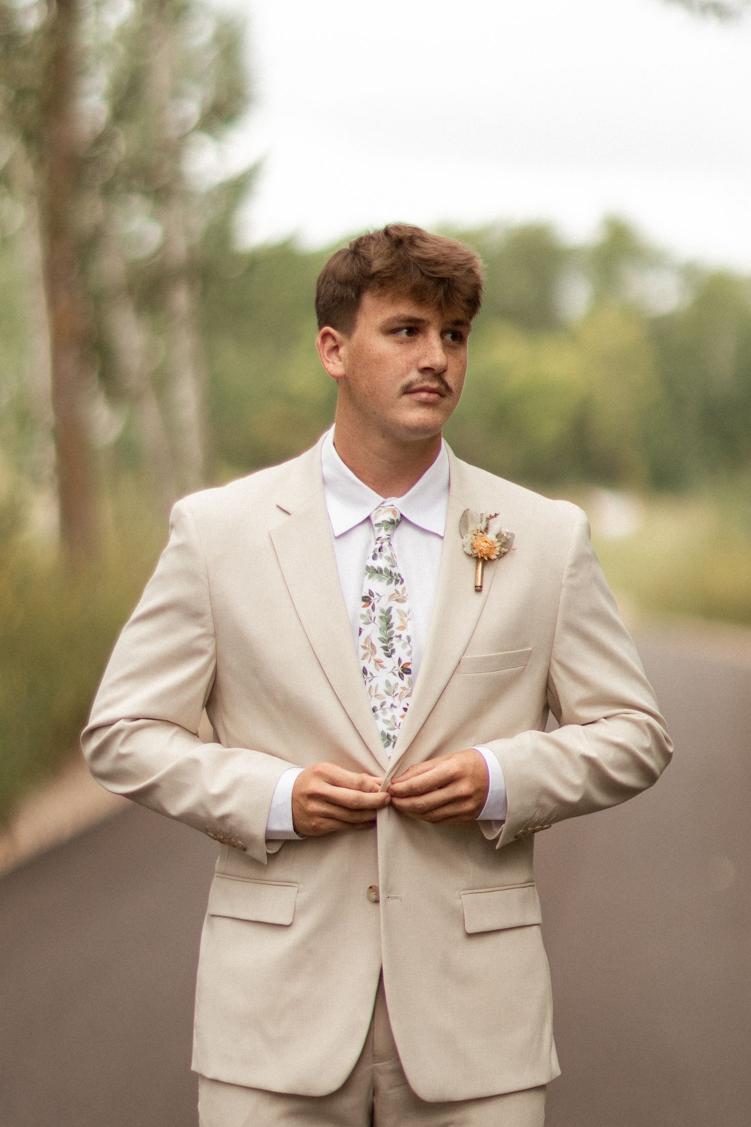 Groom in cream colored suit adjusts his tie outdoors.