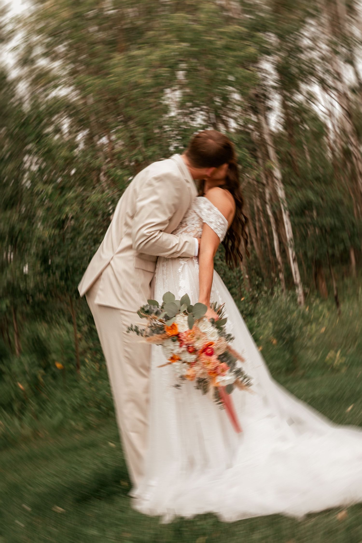 A romantic wedding couple shares an intimate kiss in a forest setting with a flowing white dress and beige suit.