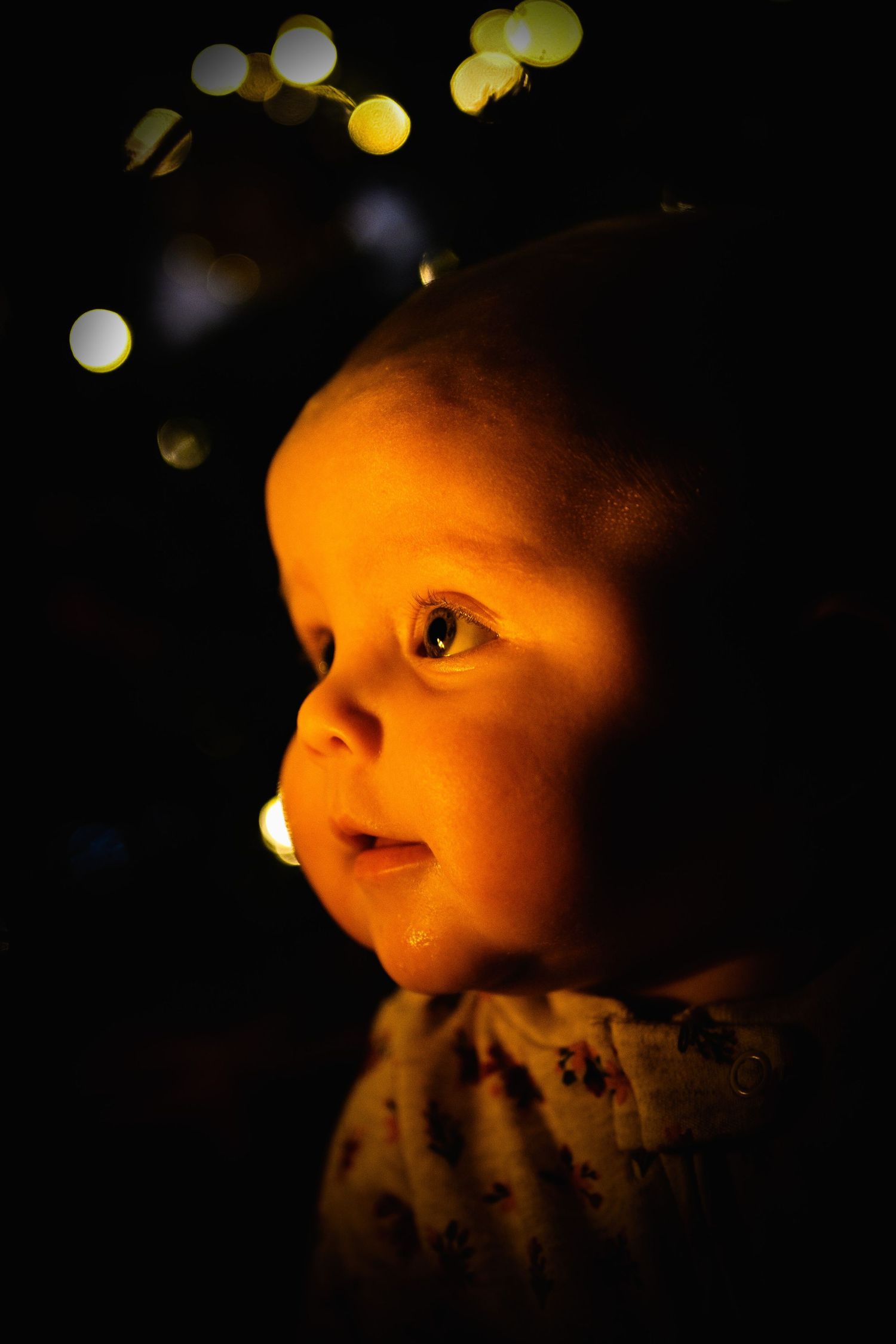 Close-up profile of baby illuminated by warm holiday lights against dark background.