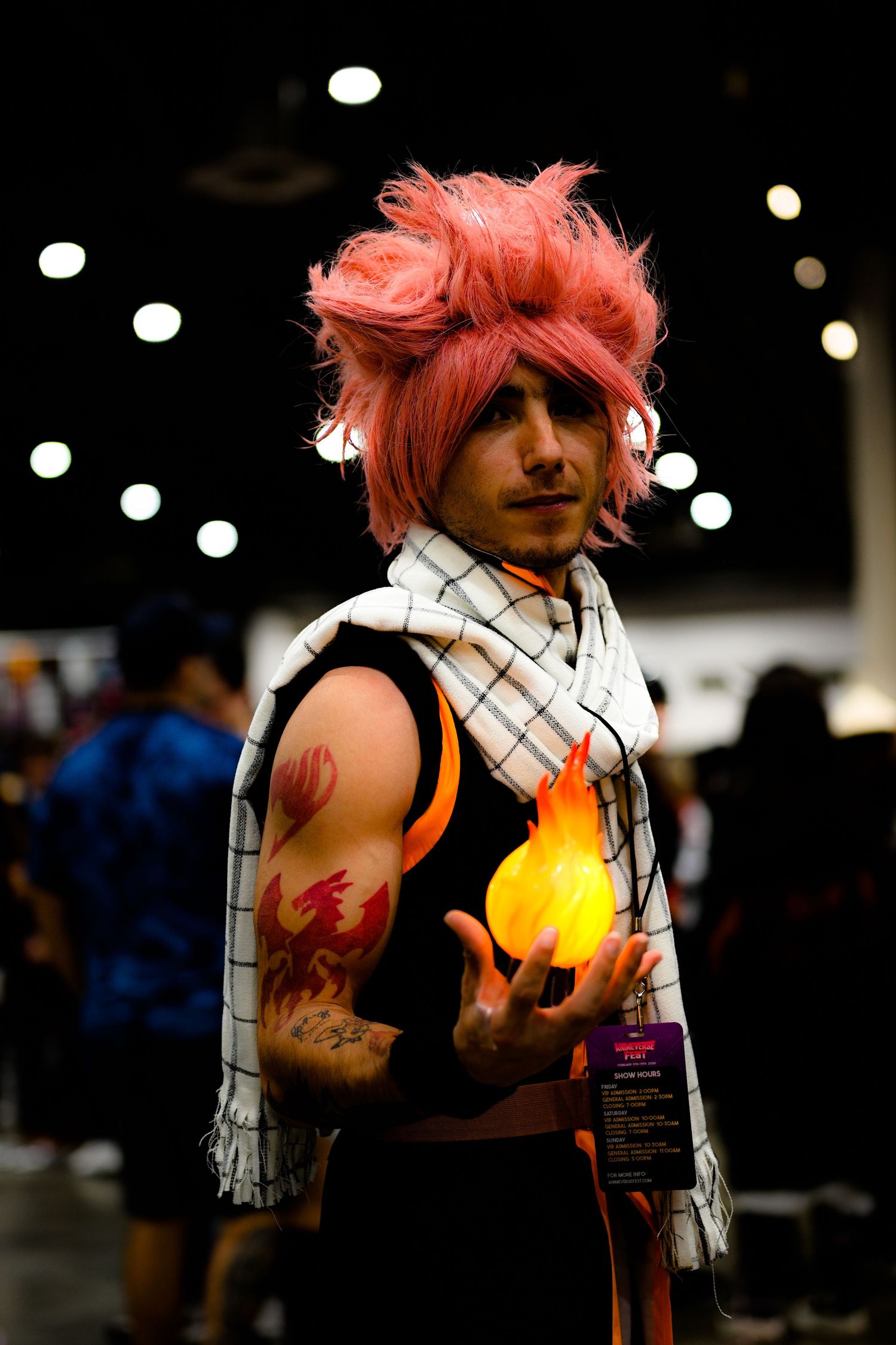 Pink-haired cosplayer in black outfit with white scarf holding glowing prop in dark setting.