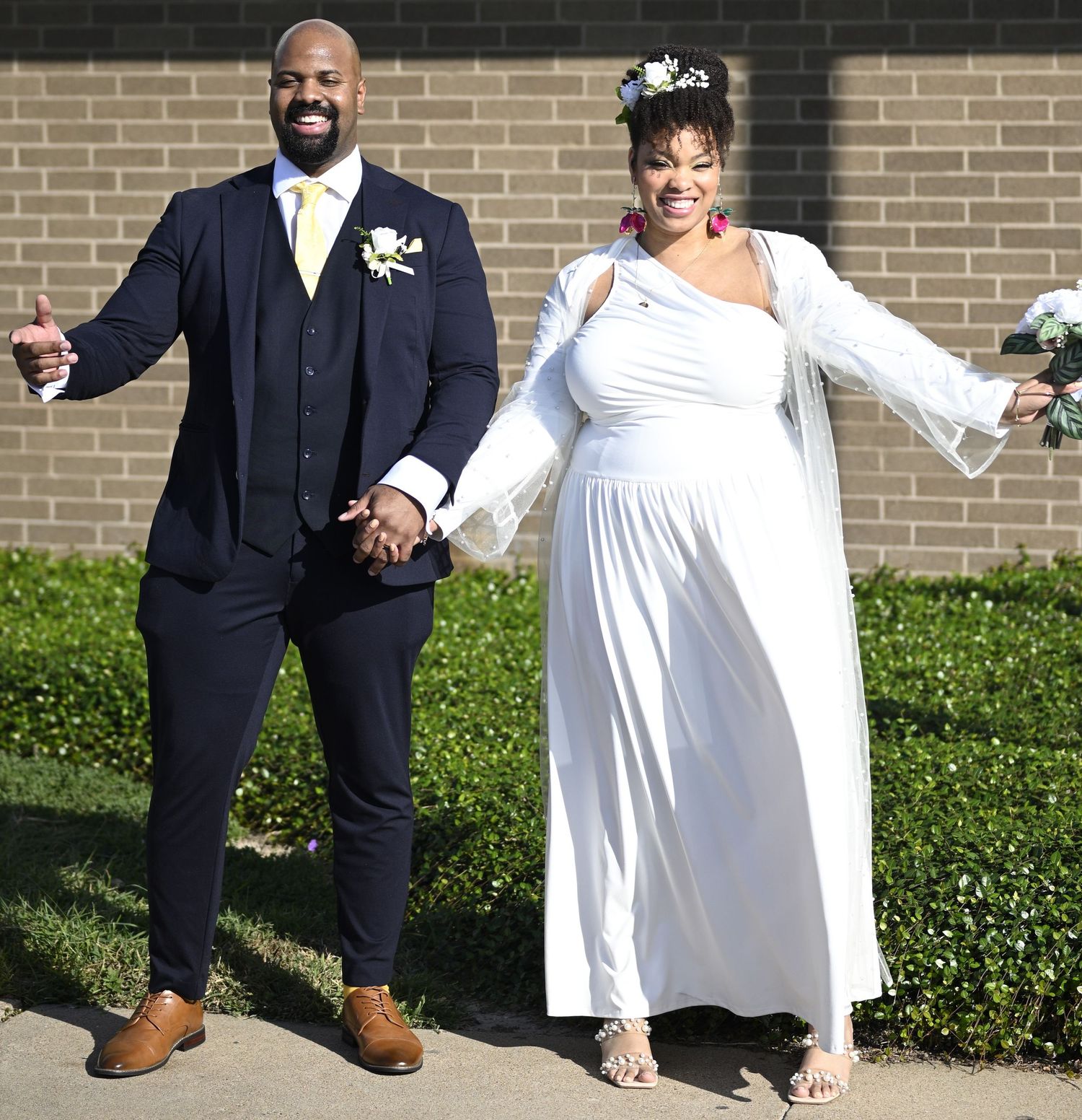 Wedding couple poses against a brick wall with greenery in formal white and navy attire.