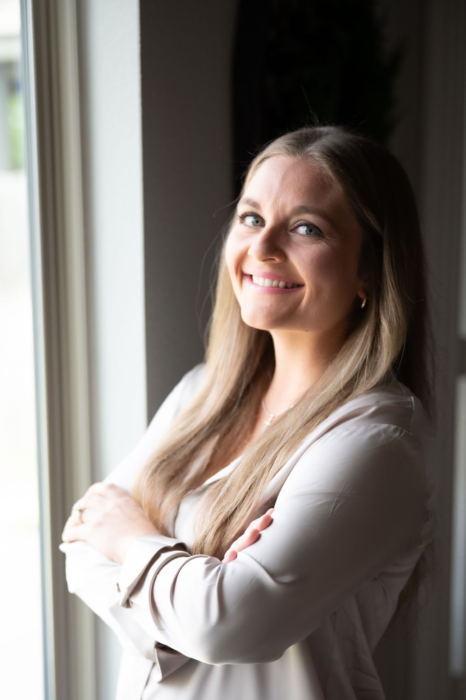 A woman in a white blouse poses by a window in natural light.