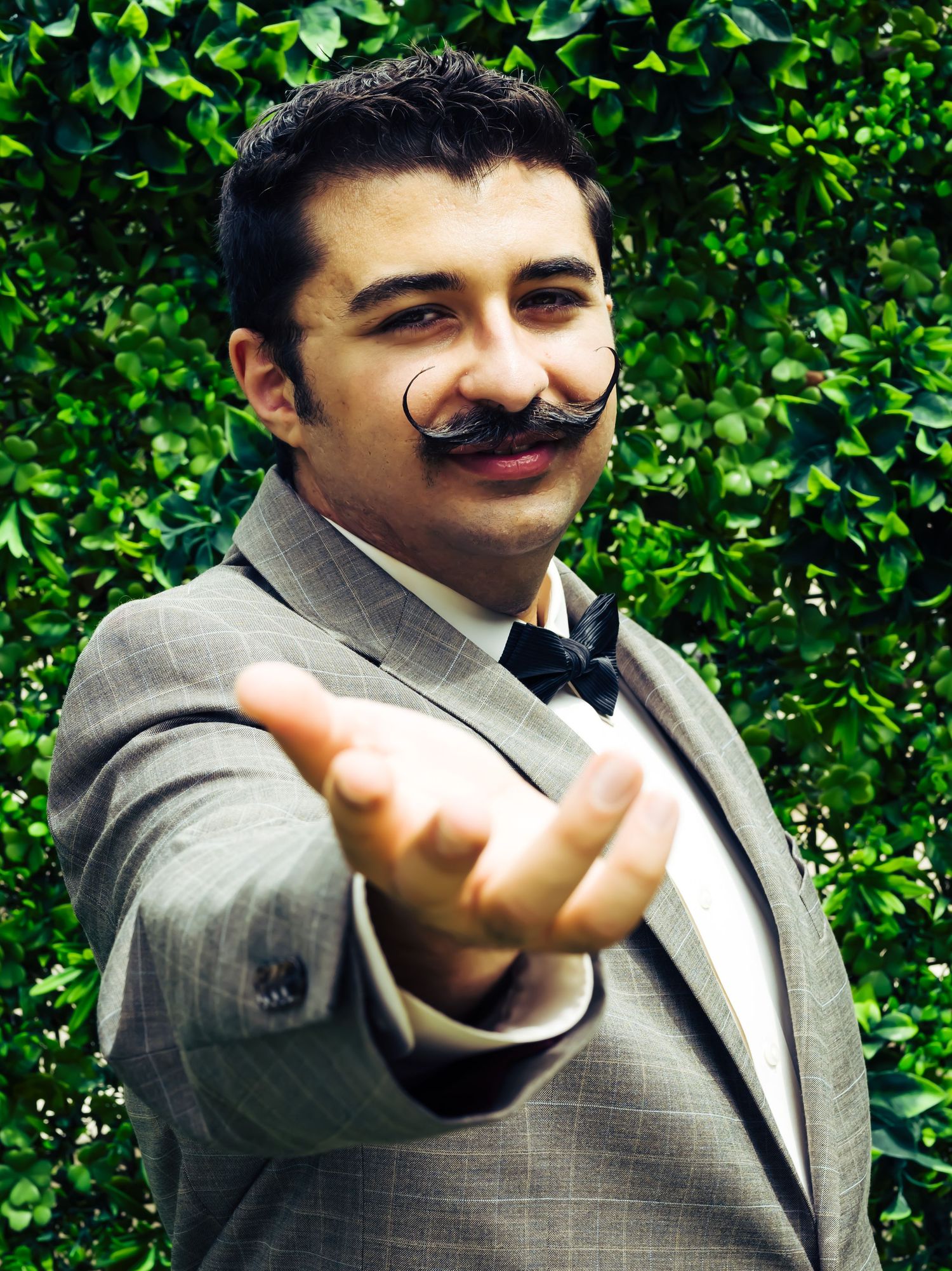 Groom in grey suit makes playful gestures against green hedge background.