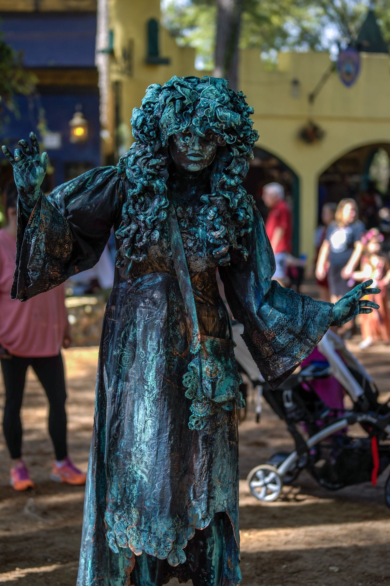 Performer in ornate teal costume and moss-like wig poses at renaissance festival.