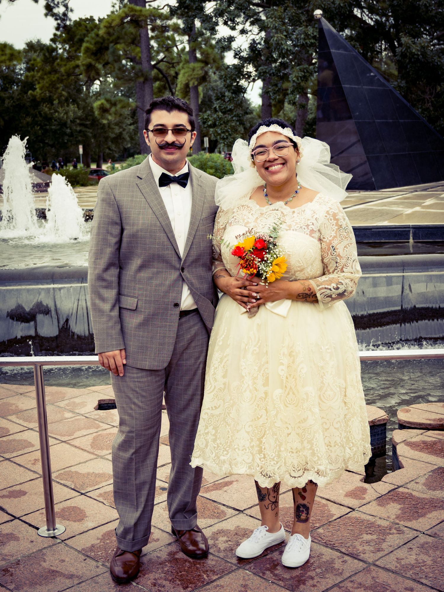 A couple in formal attire poses together on their wedding day wearing cream and grey outfits.