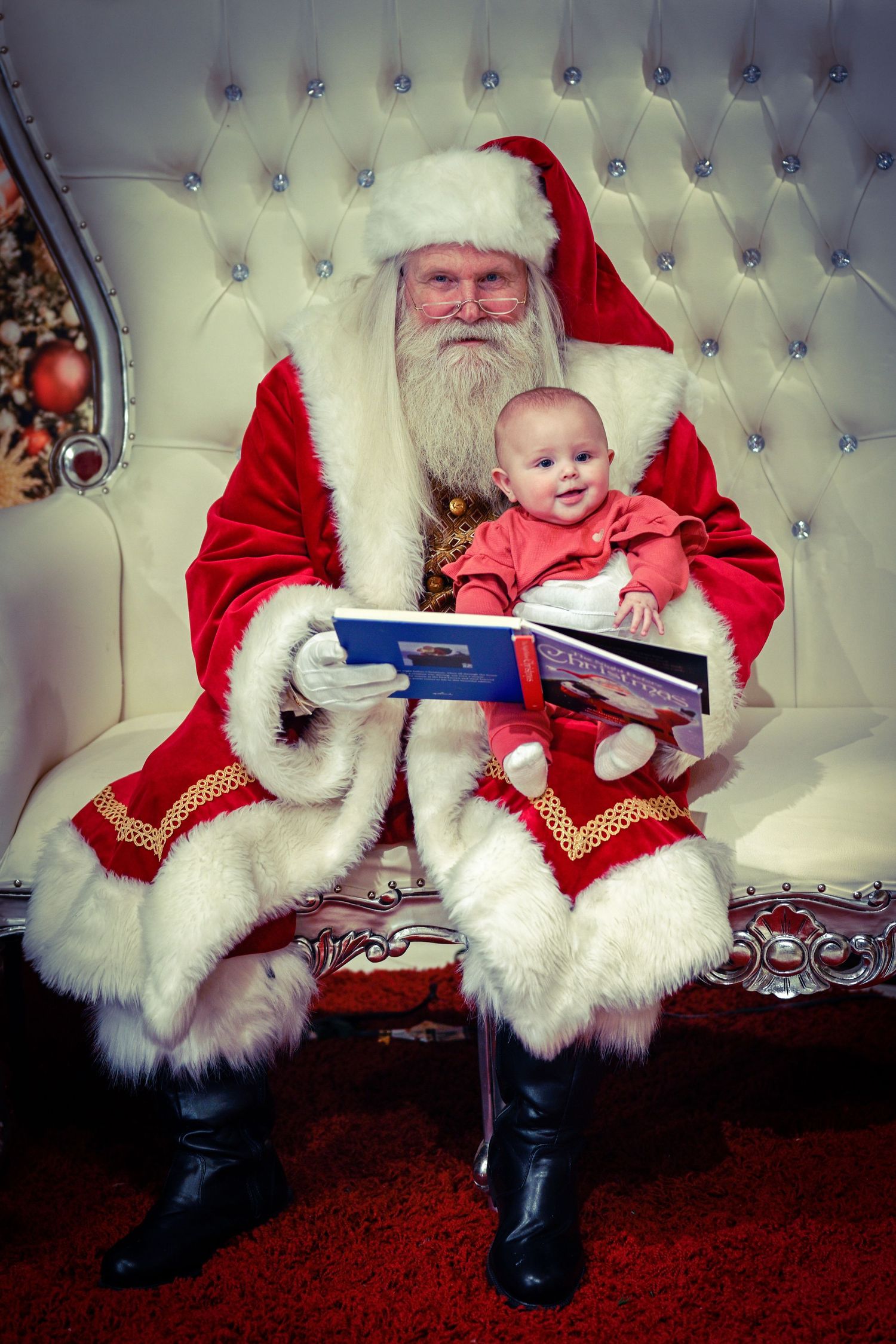 Holiday portrait of Santa reading a book to a baby on traditional white throne setting.