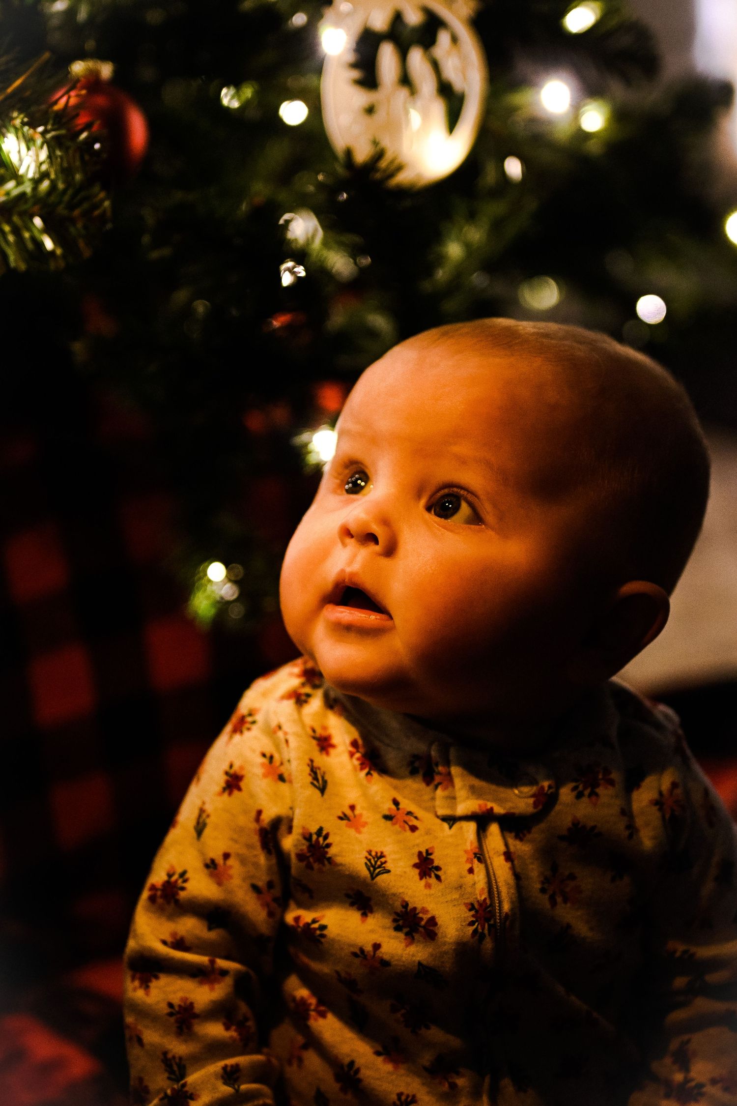 Baby looking up at Christmas tree lights with amazement in dark room.