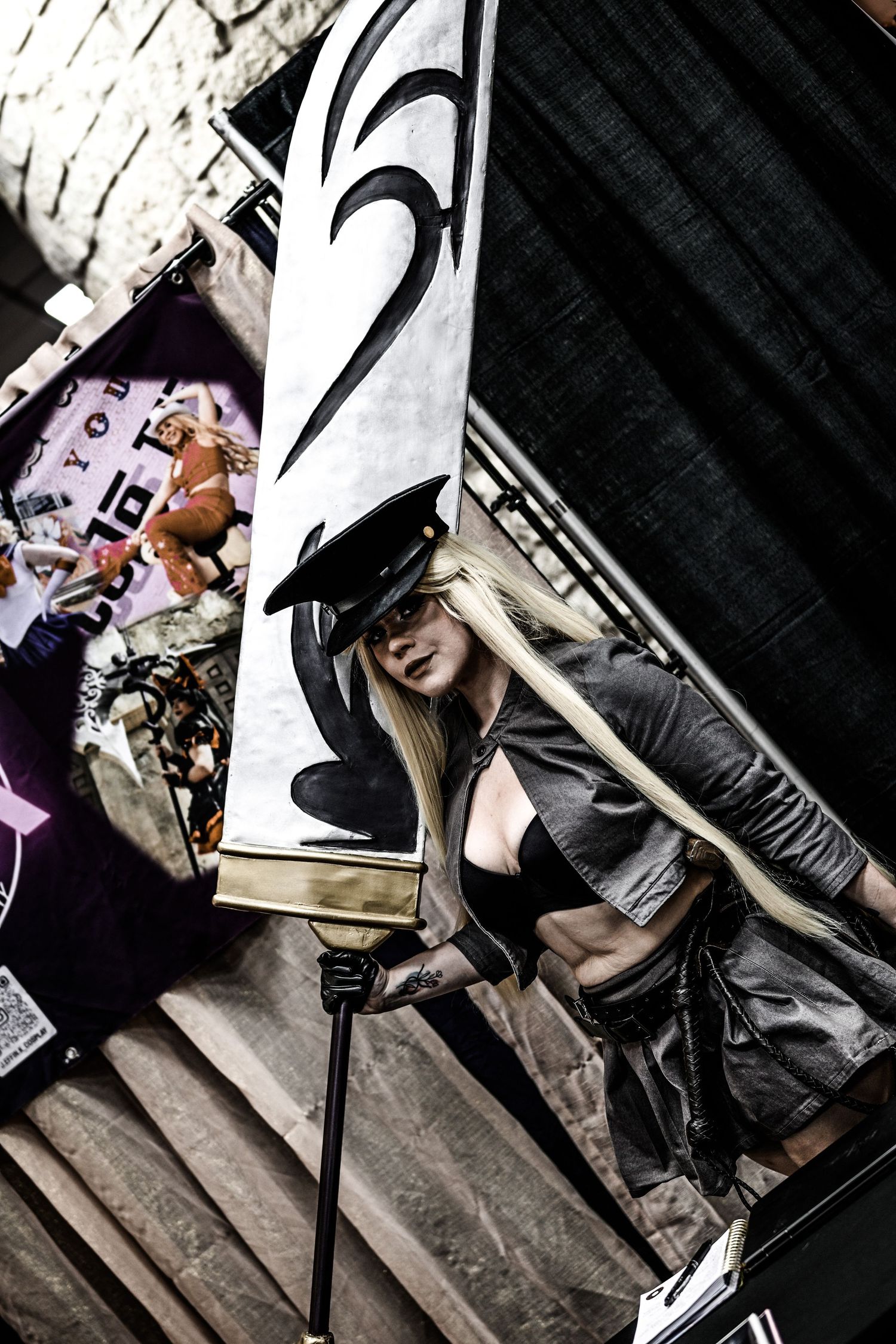 Cosplayer in dark costume and hat posing with large banner at convention.