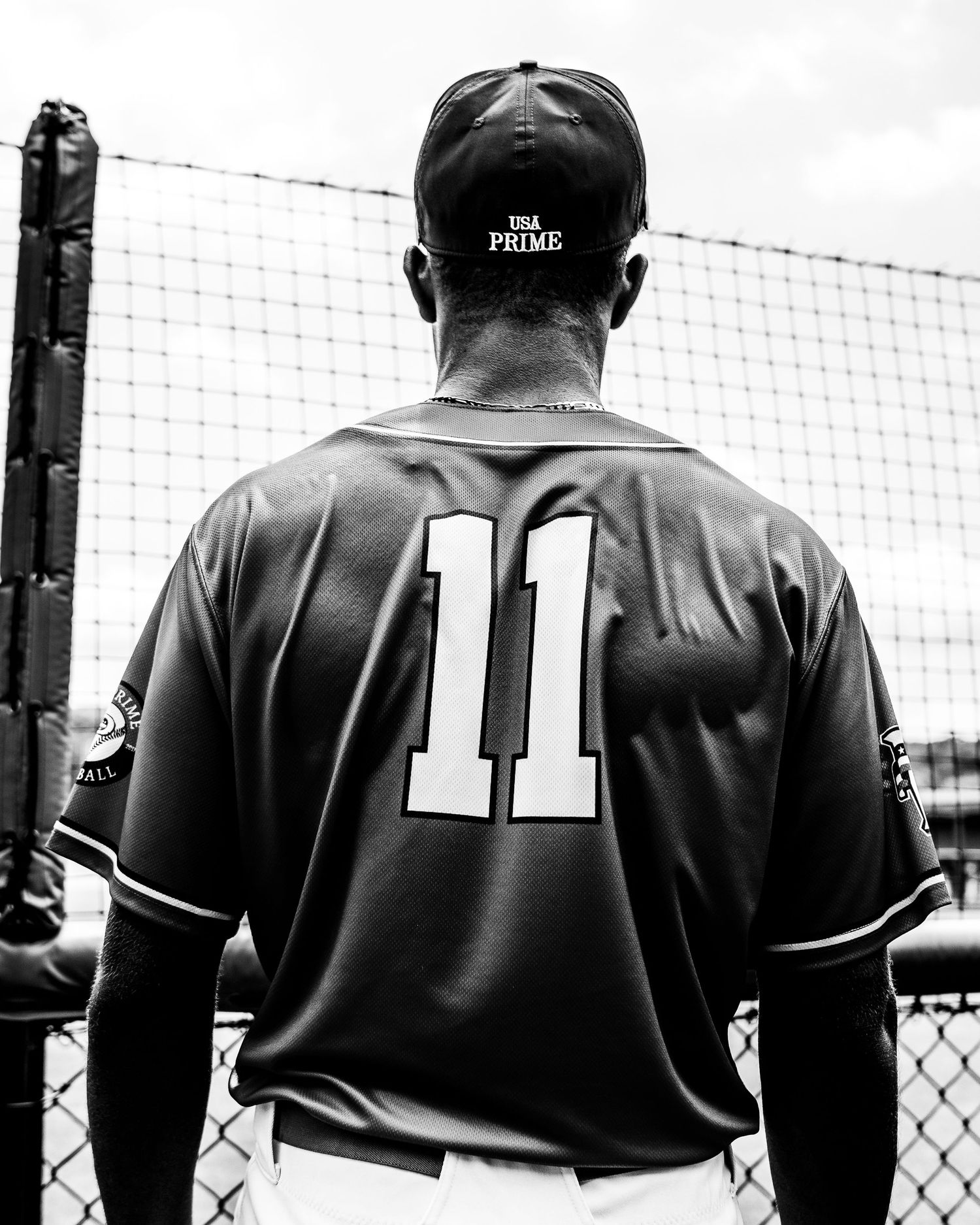Black and white photo of baseball player wearing USA Prime jersey number 11, viewed from behind near batting cage.