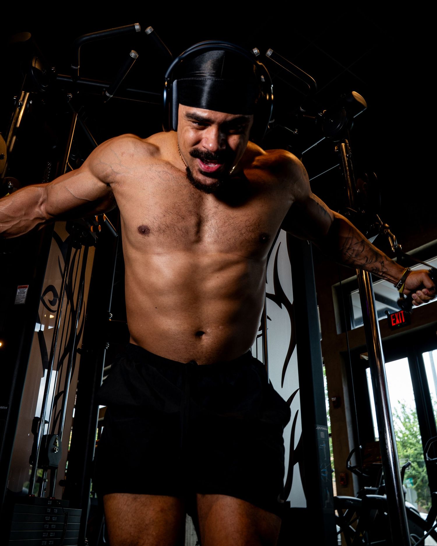 Muscular shirtless man performing cable fly exercise on gym machine, showcasing defined abs and arms.
