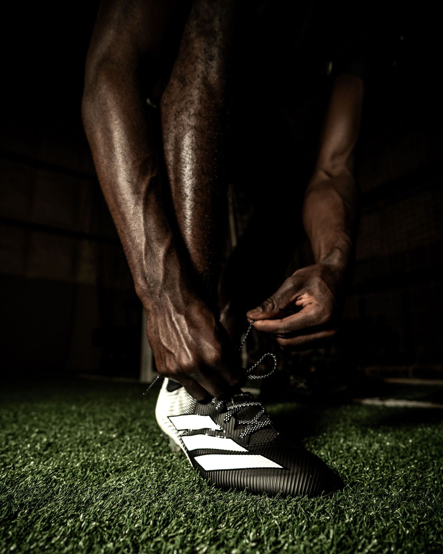 Athlete tying Adidas cleats on green turf in dramatic lighting, showcasing athletic preparation and sportswear.