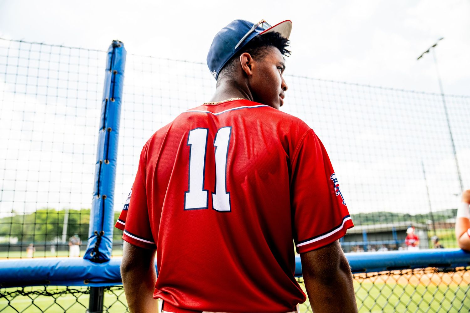 Baseball player wearing red jersey number 11 stands near a chain-link fence on a sunny day at a baseball field.