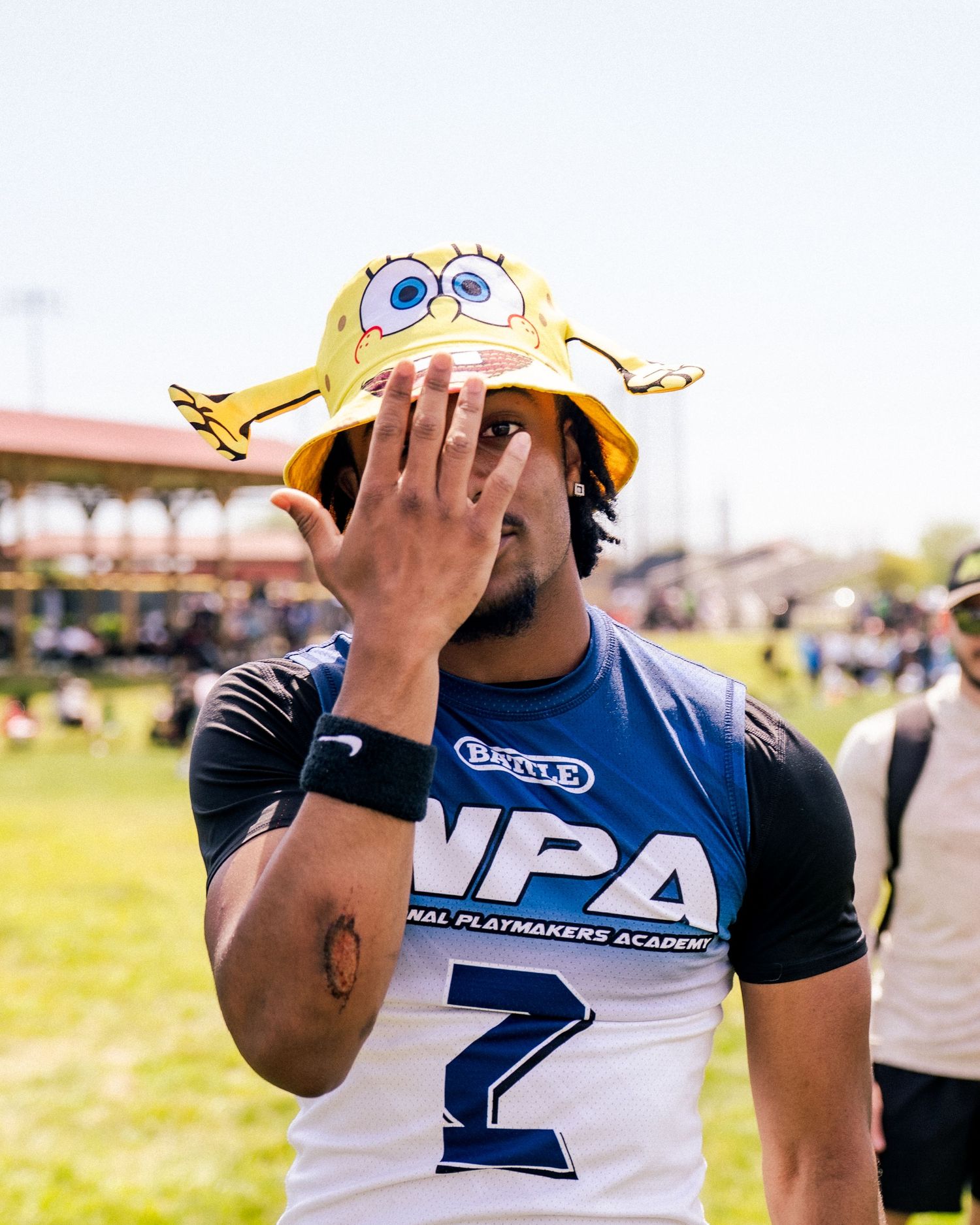 Athlete wearing SpongeBob bucket hat and NPA jersey number 2 covers face with hand at outdoor sports event.