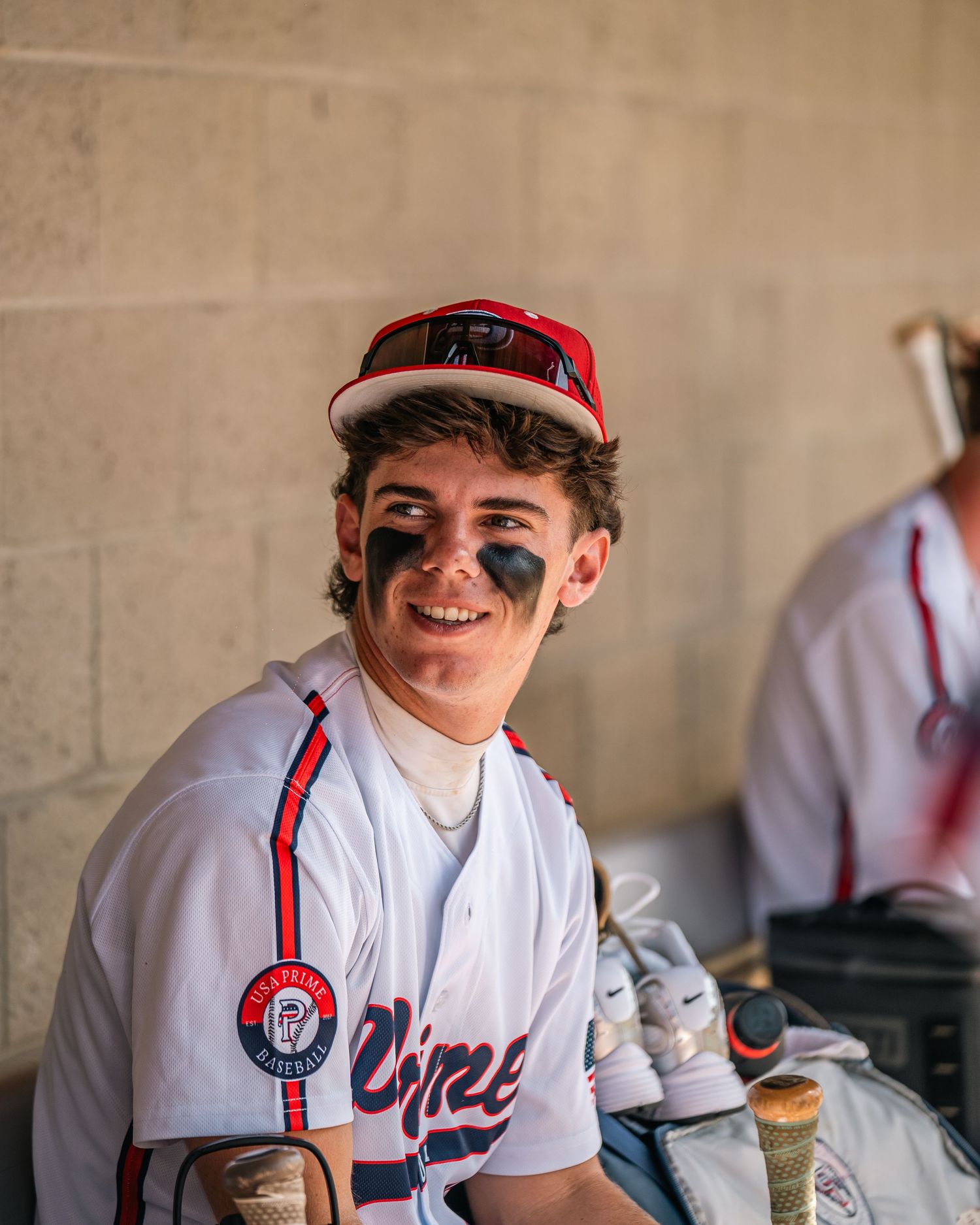 Smiling baseball player in dugout wearing white uniform with eye black and red cap holding bats.