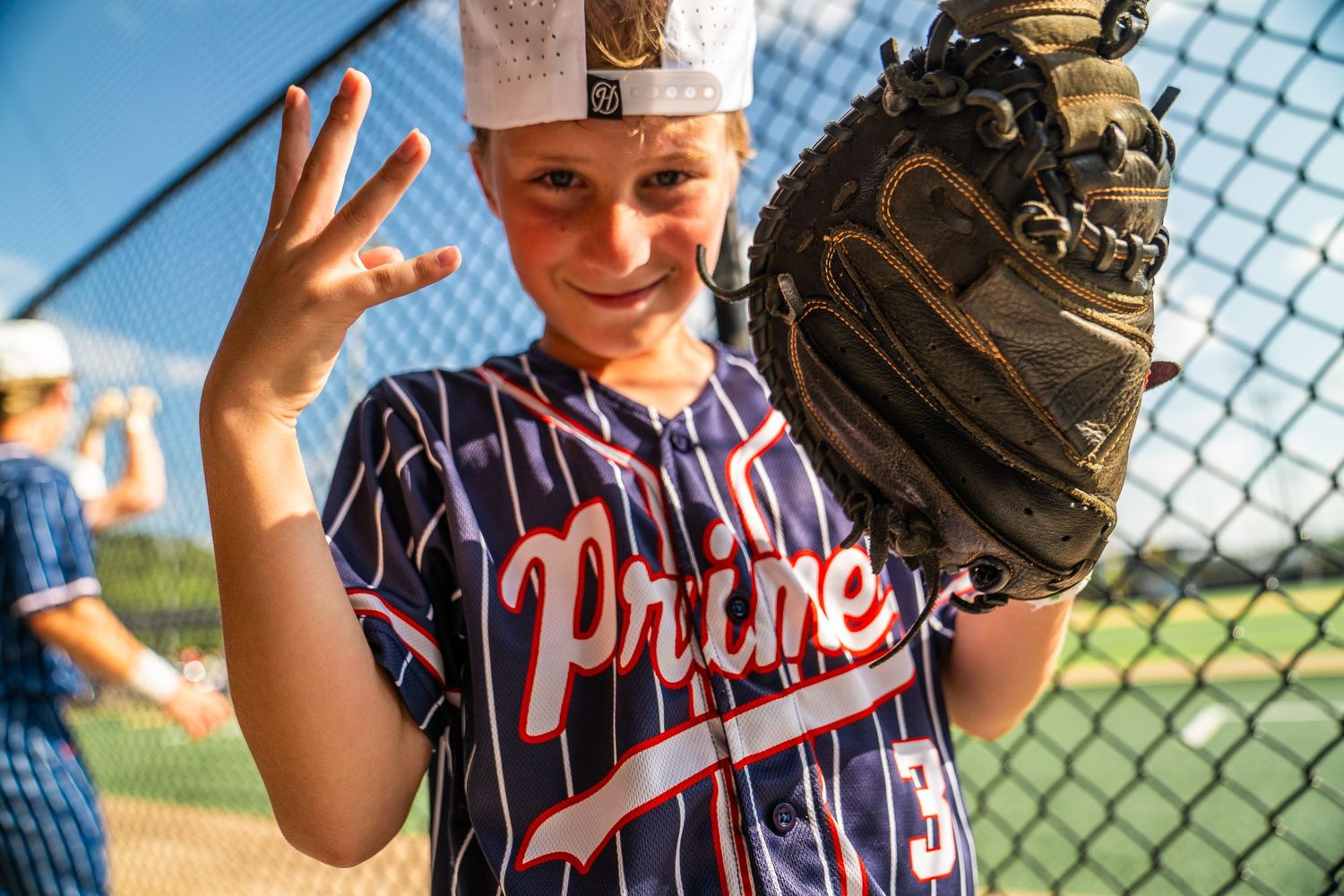 Young baseball player in Prime jersey holds up glove and fingers near dugout fence on sunny day.