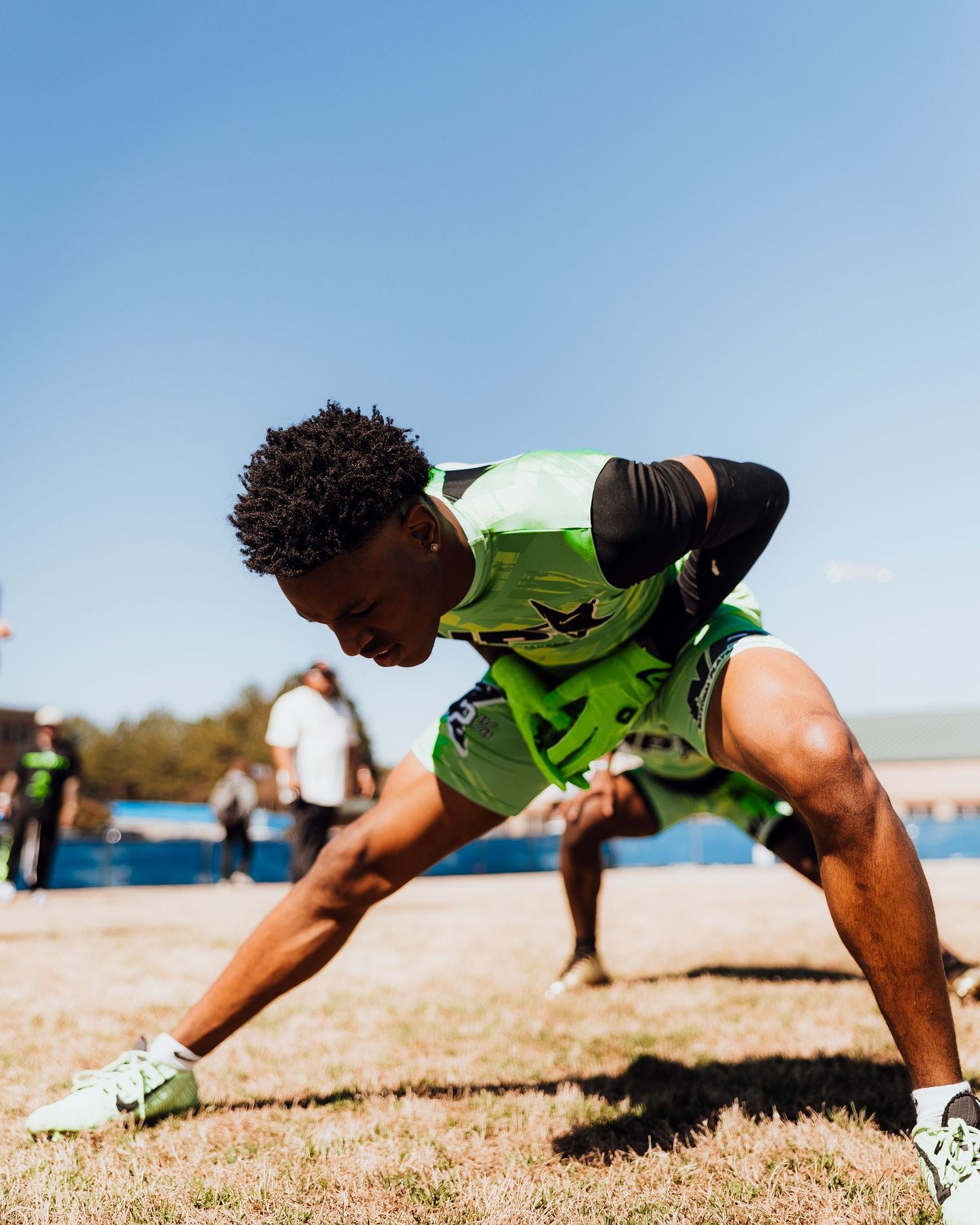 Young athlete in green uniform crouches in starting position on grass field under clear blue sky.